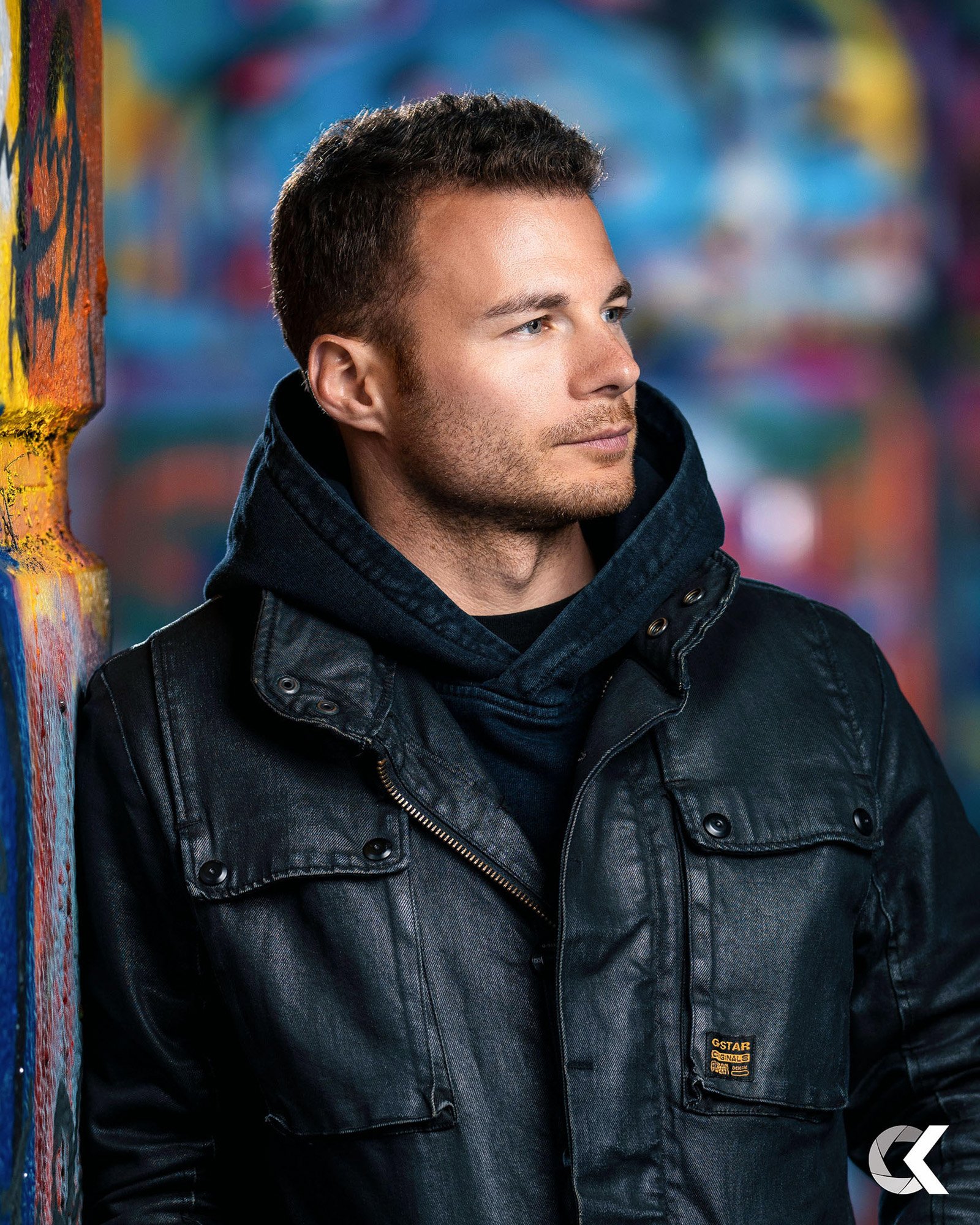 A young man with short brown hair and light stubble looking thoughtfully to the right, wearing a black leather jacket and a dark hoodie against a colorful graffiti background.