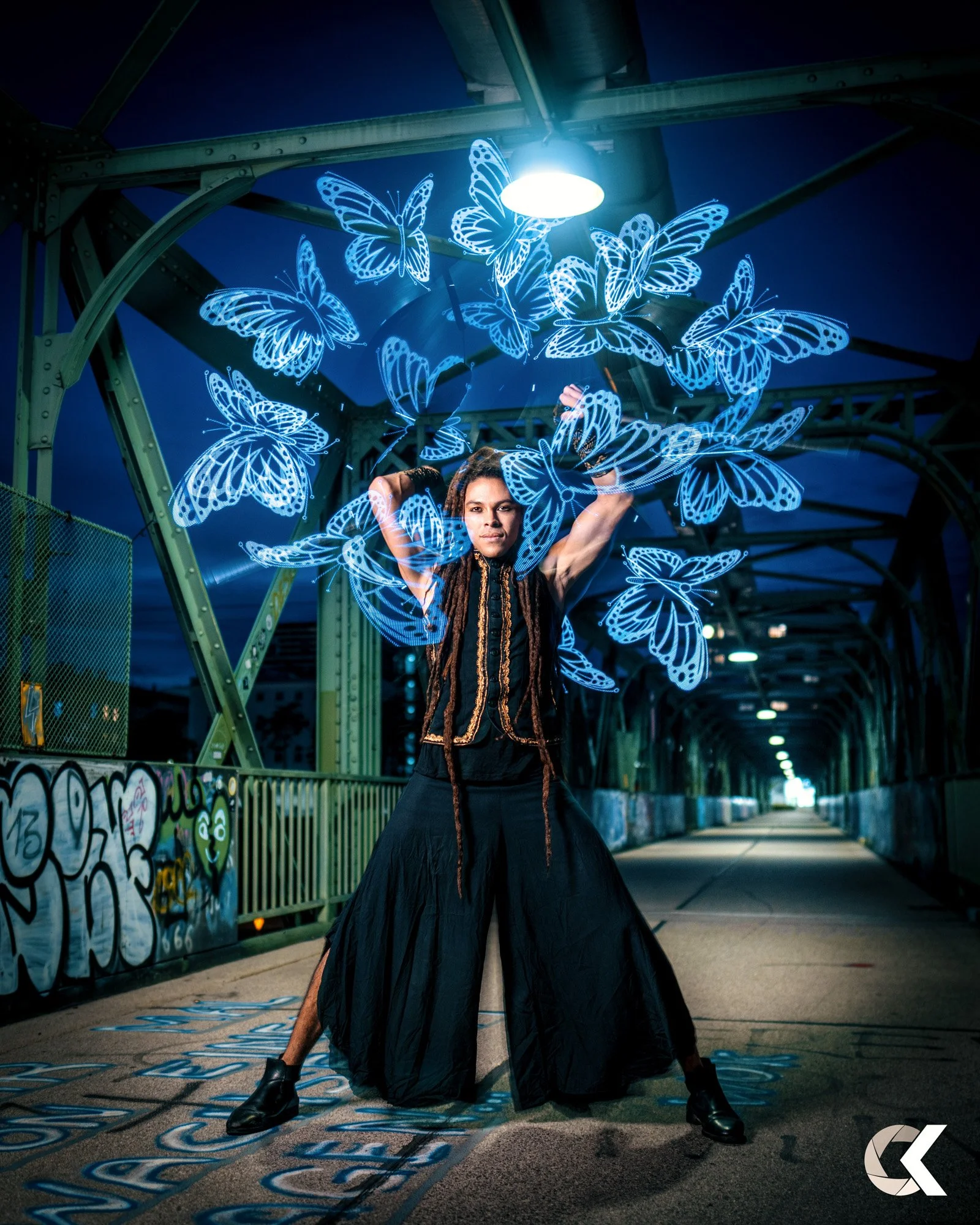 A person standing on a bridge with graffiti, holding neon blue butterfly props that glow and surround them, under a mounted light at night.