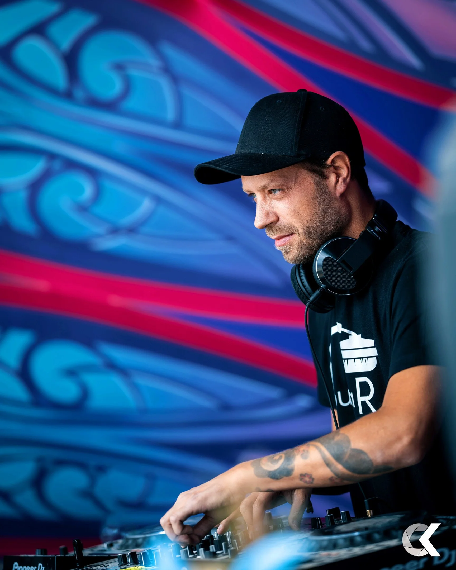 A DJ wearing a black cap and headphones, DJing against a colorful, abstract background with blue, red, and pink swirls.