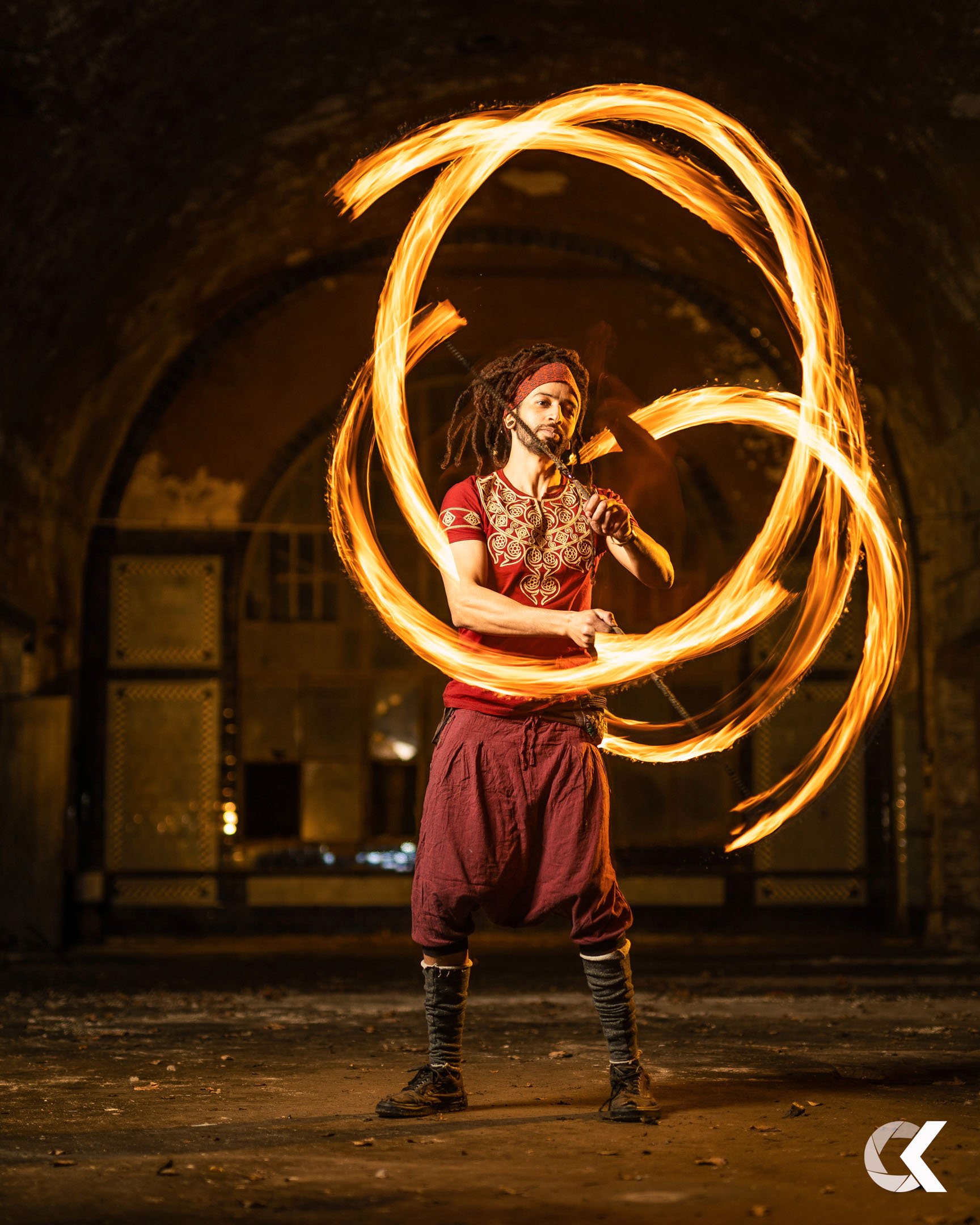 A man with dreadlocks and a red headband performing a fire spinning act in a dimly lit space, creating circular trails of fire around him.