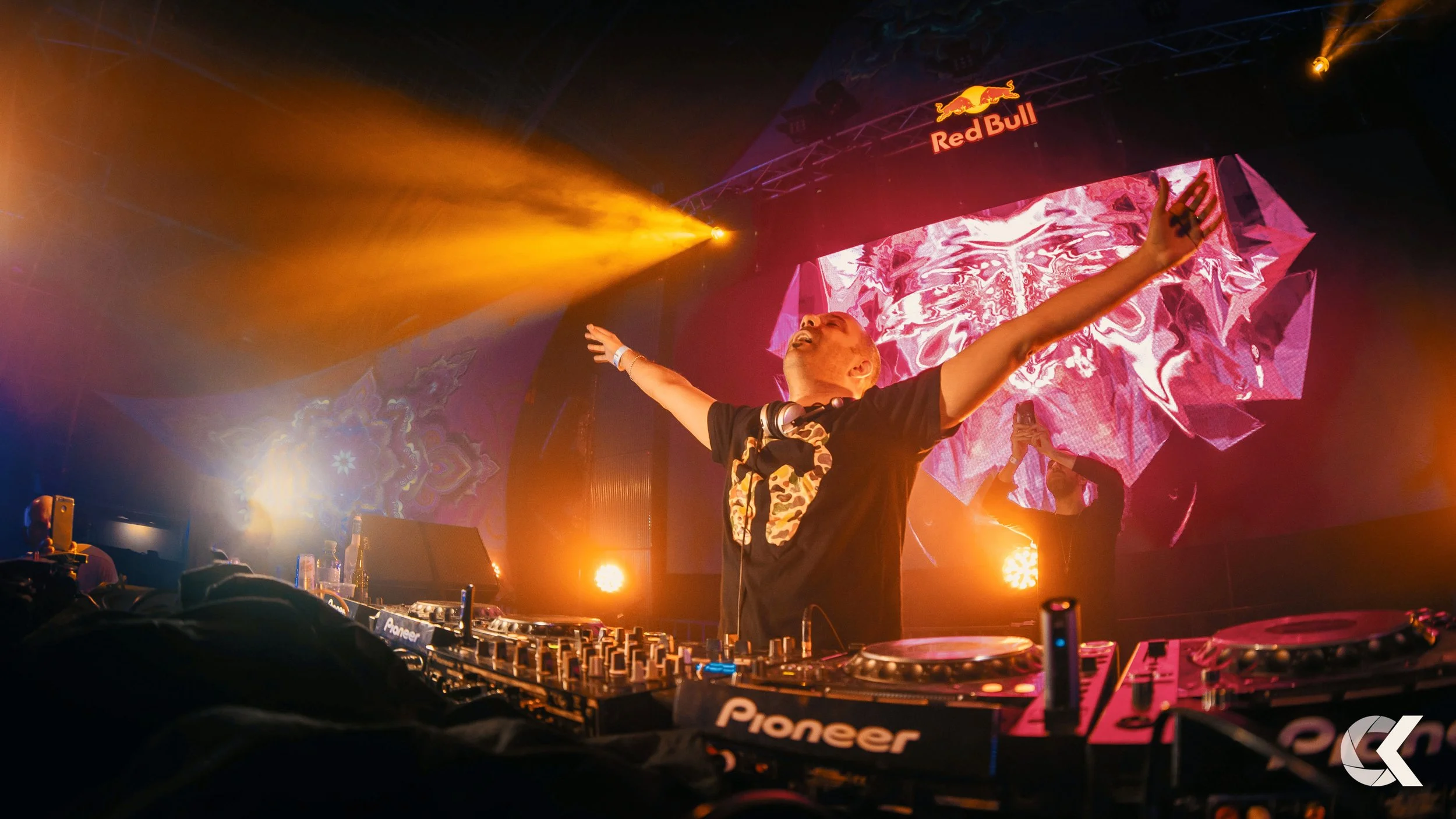 DJ performing at a Red Bull event with arms raised, colorful abstract background, and bright stage lights.
