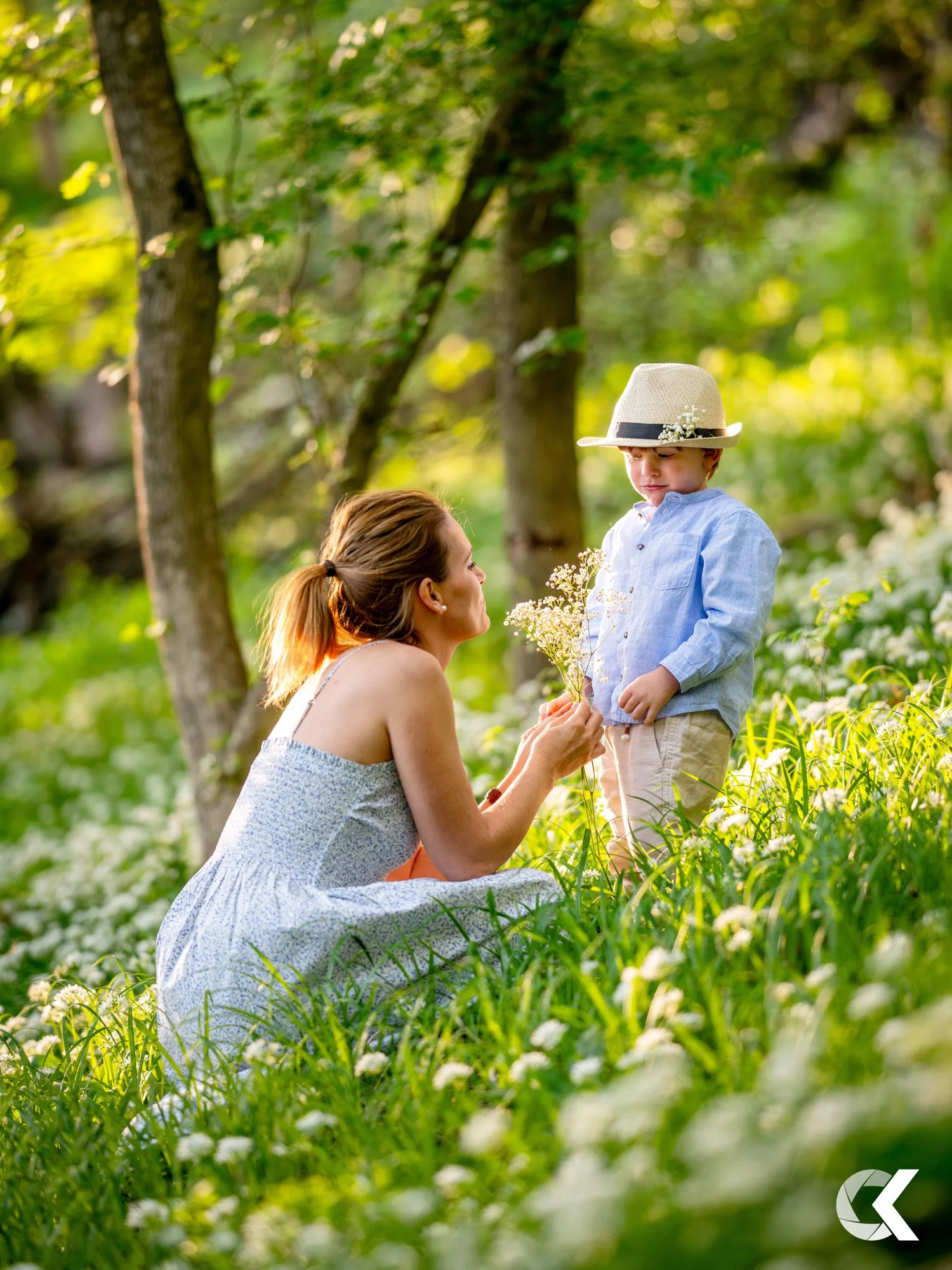 A woman kneeling in a green field with white flowers, holding a small bouquet, looking at a young boy in a blue shirt and beige shorts who is standing in front of her, wearing a hat with a black band and white decoration, holding a small bouquet of w