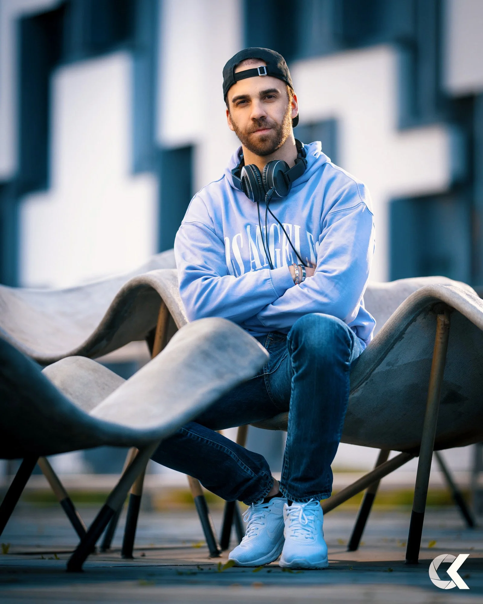 A young man wearing a black backwards cap, a light blue hoodie, and jeans, sitting outdoors on a curved modern bench, with headphones around his neck and arms crossed.