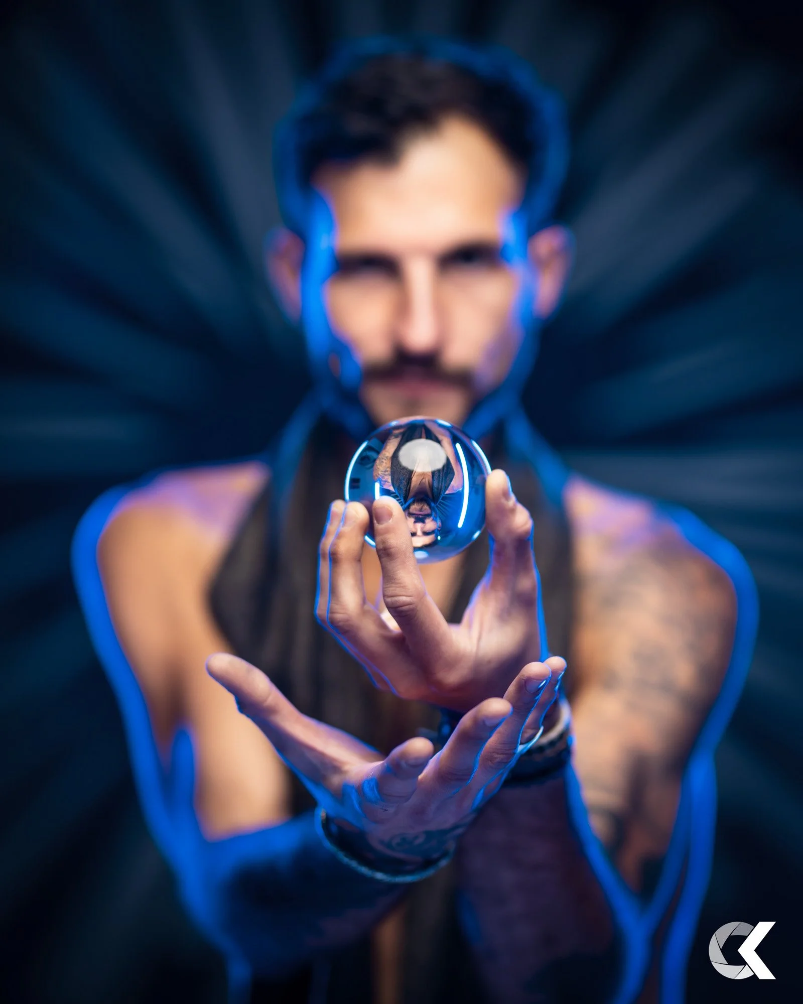 A man with dark hair and a mustache, wearing a sleeveless shirt, holds a reflective sphere with a cityscape image inside. The background features dynamic streaks of light, creating a dramatic effect.