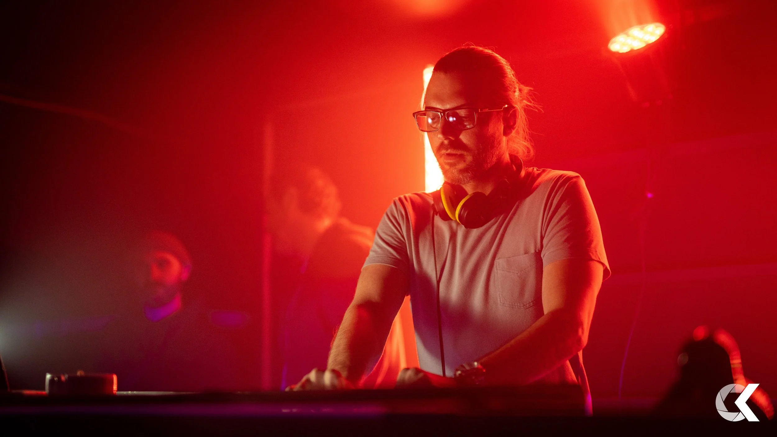 A DJ wearing glasses and headphones around his neck, working at a turntable in a red-lit club environment with two other people in the background.