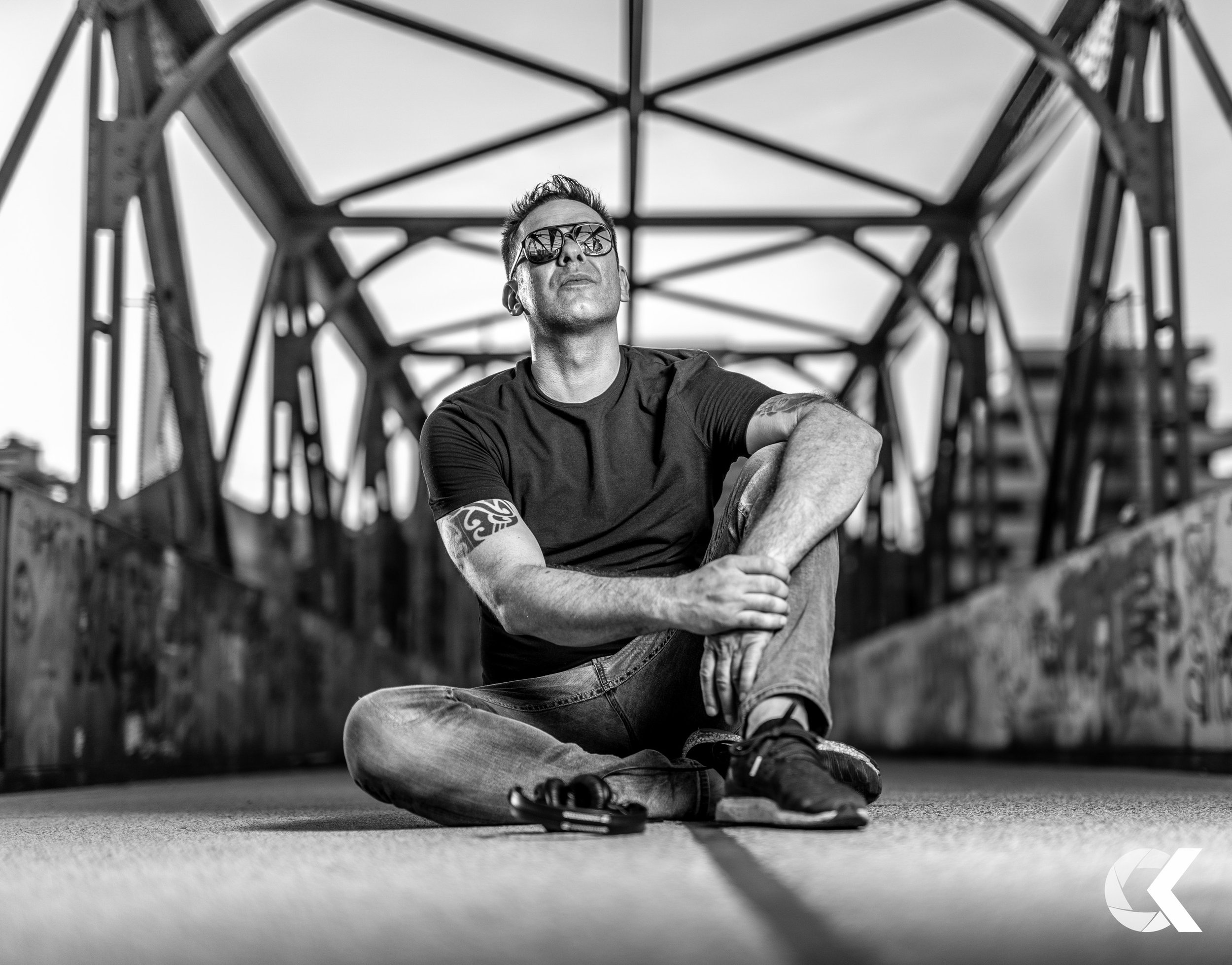 A man wearing sunglasses sits cross-legged on the ground underneath a bridge with steel beams, looking up. The image is in black and white.
