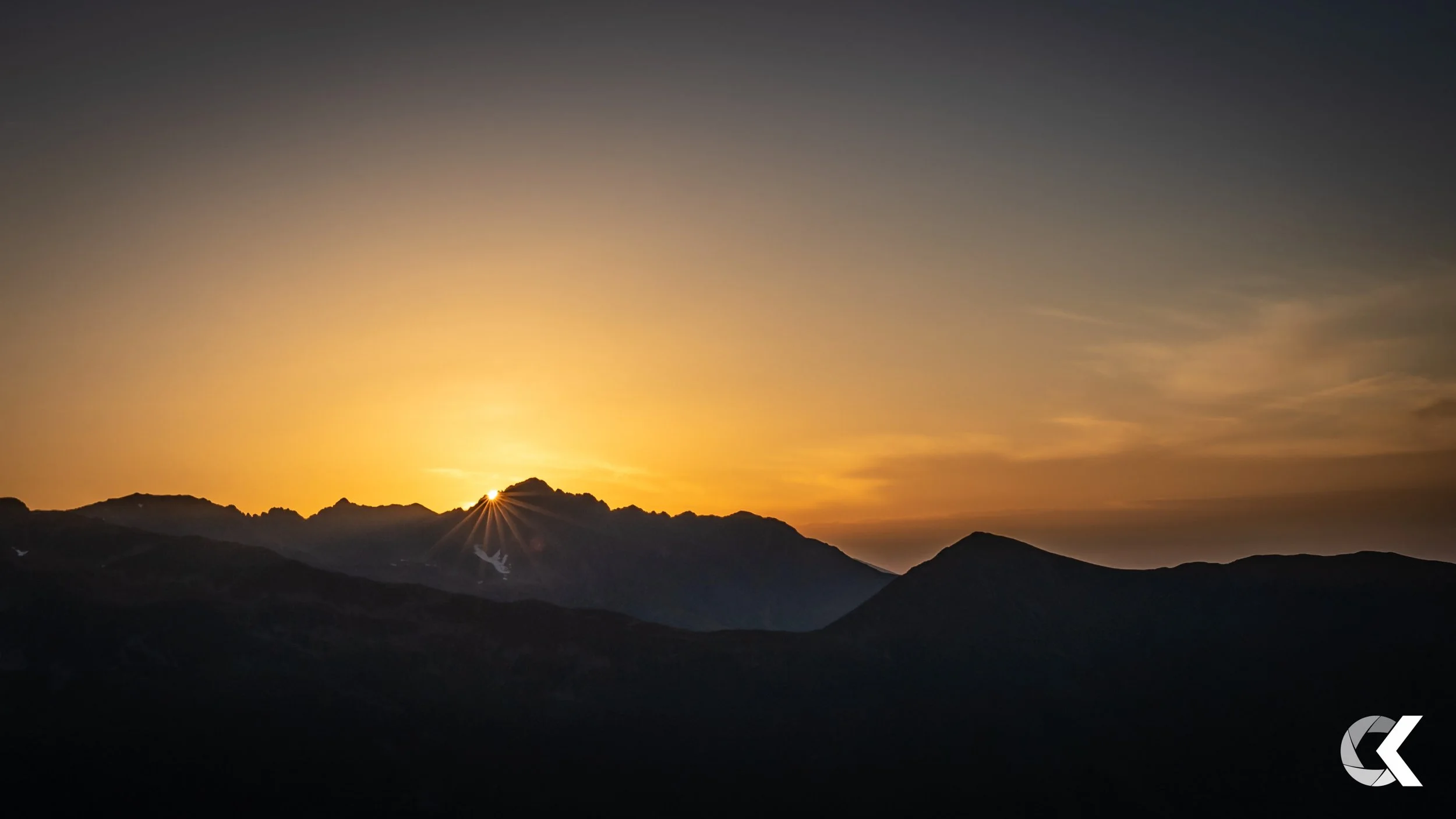 Sunset over mountain range with the sun partially hidden behind jagged peaks, casting a warm glow in the sky.