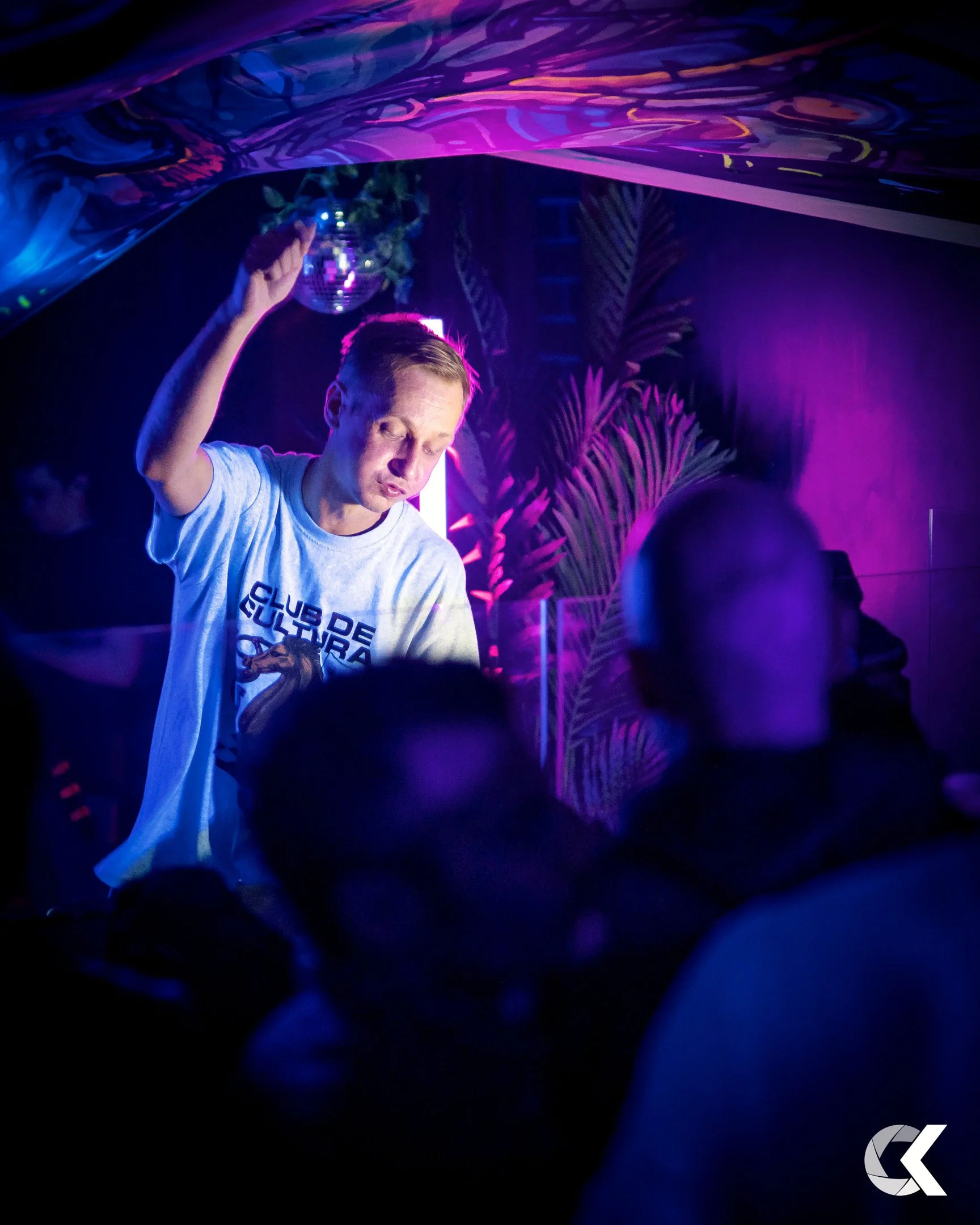A man dancing at a nightclub with purple and blue lighting, wearing a white T-shirt that says 'Club de Cultura' and surrounded by other people.