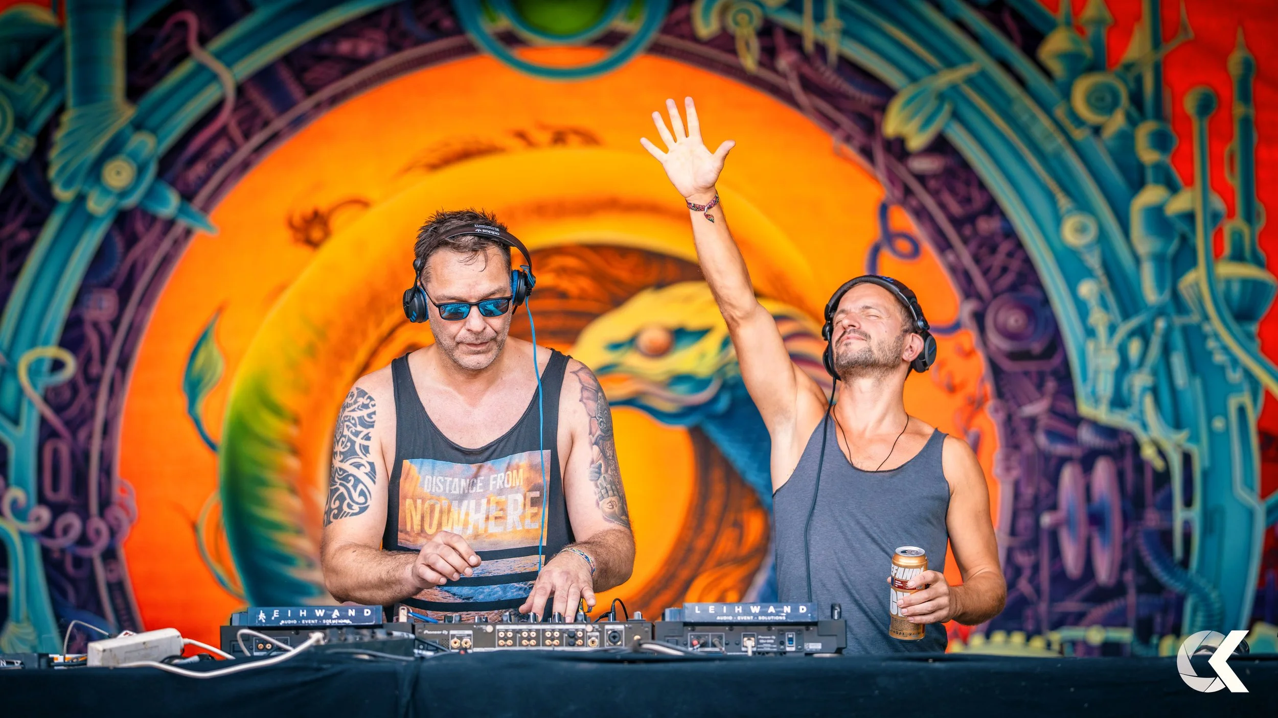 Two men DJing on stage at an electronic music event, with colorful digital art background and one holding a beverage can.