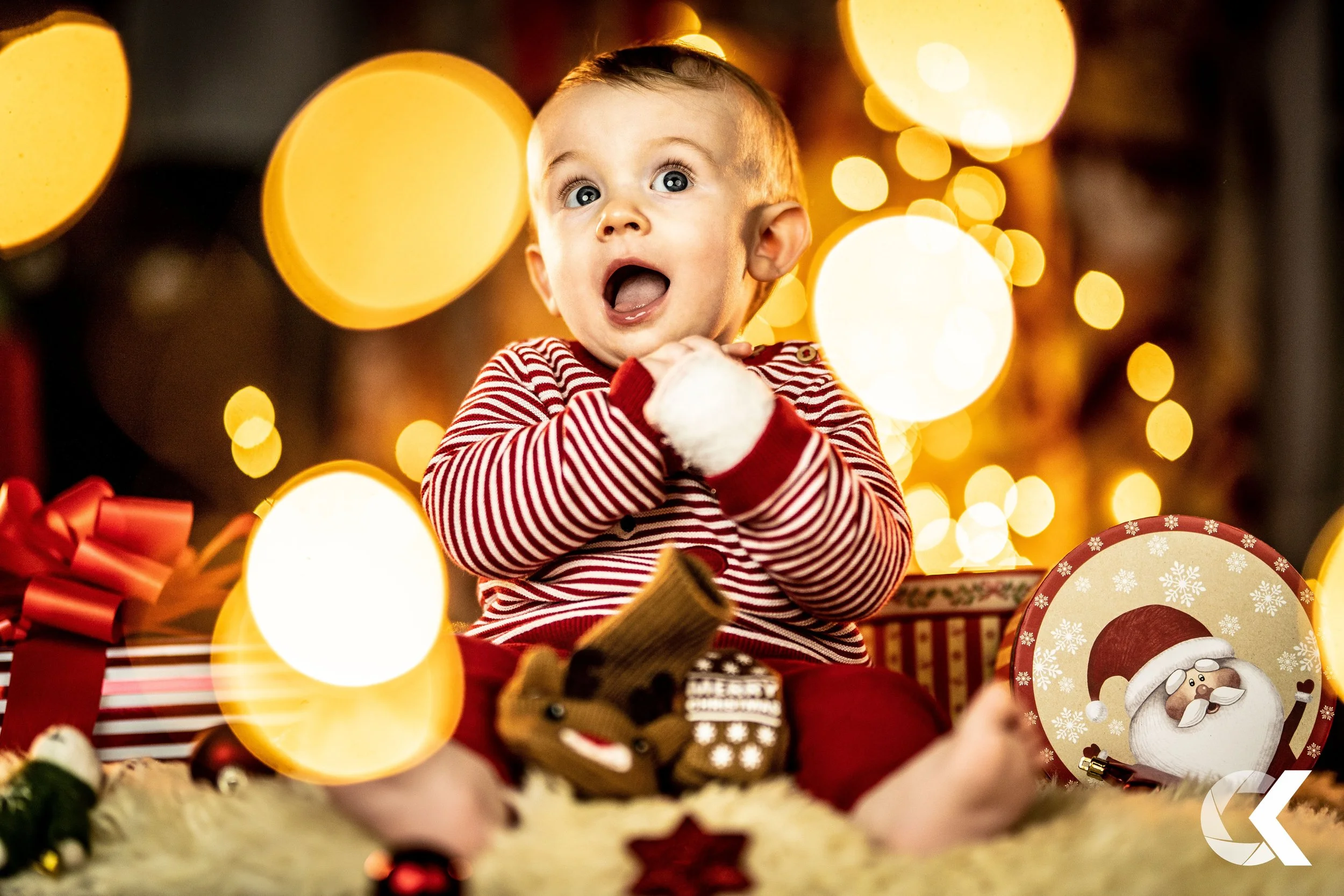 A surprised baby sitting on the floor during Christmas, surrounded by presents, decorations, and a Santa face plate, with warm yellow holiday lights in the background.