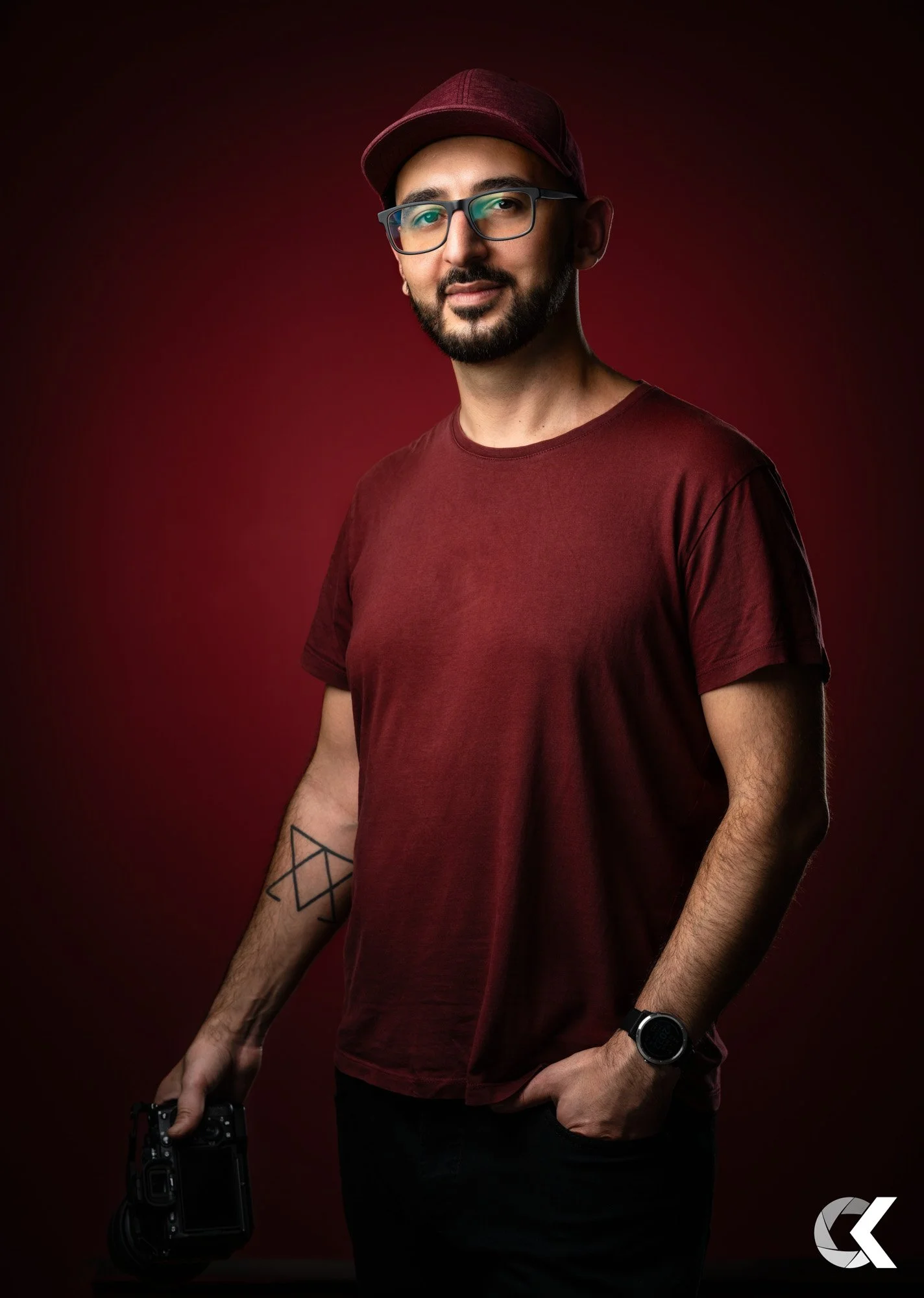A man with glasses and a beard, wearing a maroon cap and matching t-shirt, holding a camera in his right hand, standing against a dark red background.