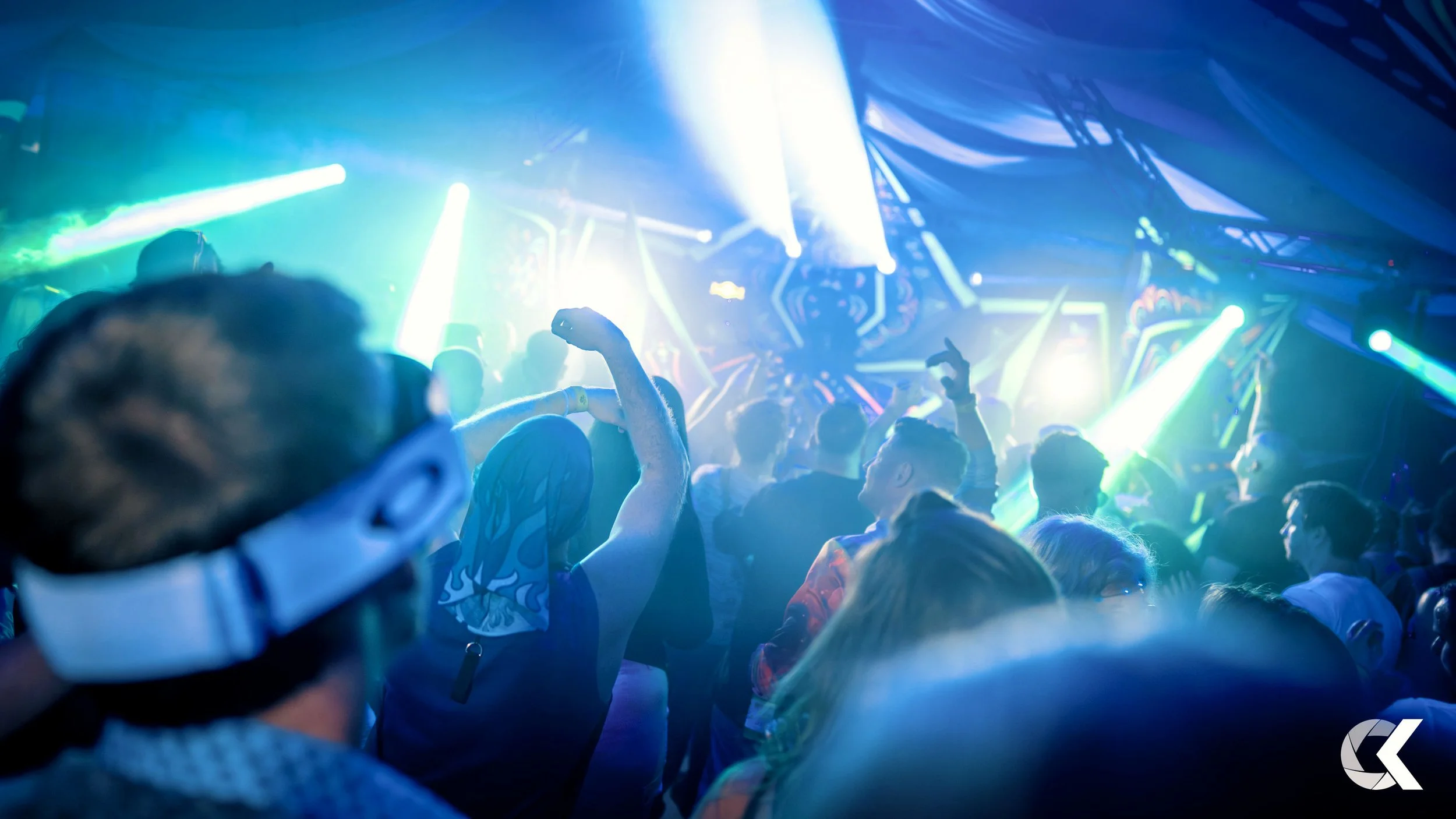 People dancing and enjoying a concert or music event under colorful stage lighting with laser effects.