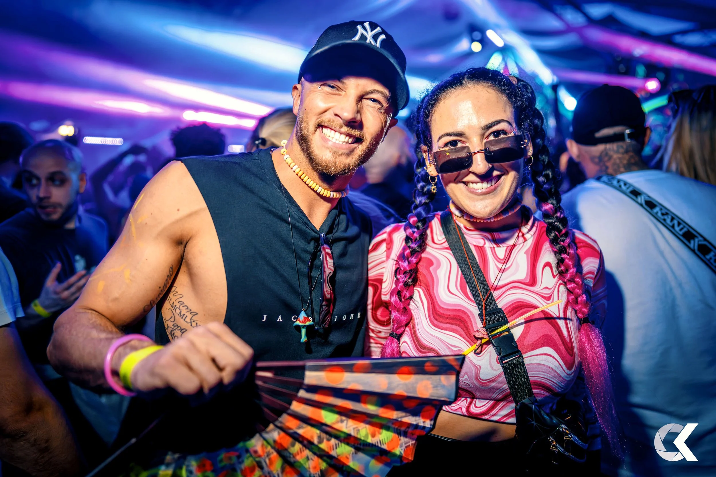 Two people smiling at a party, one holding a colorful fan, surrounded by a lively crowd and neon lights.