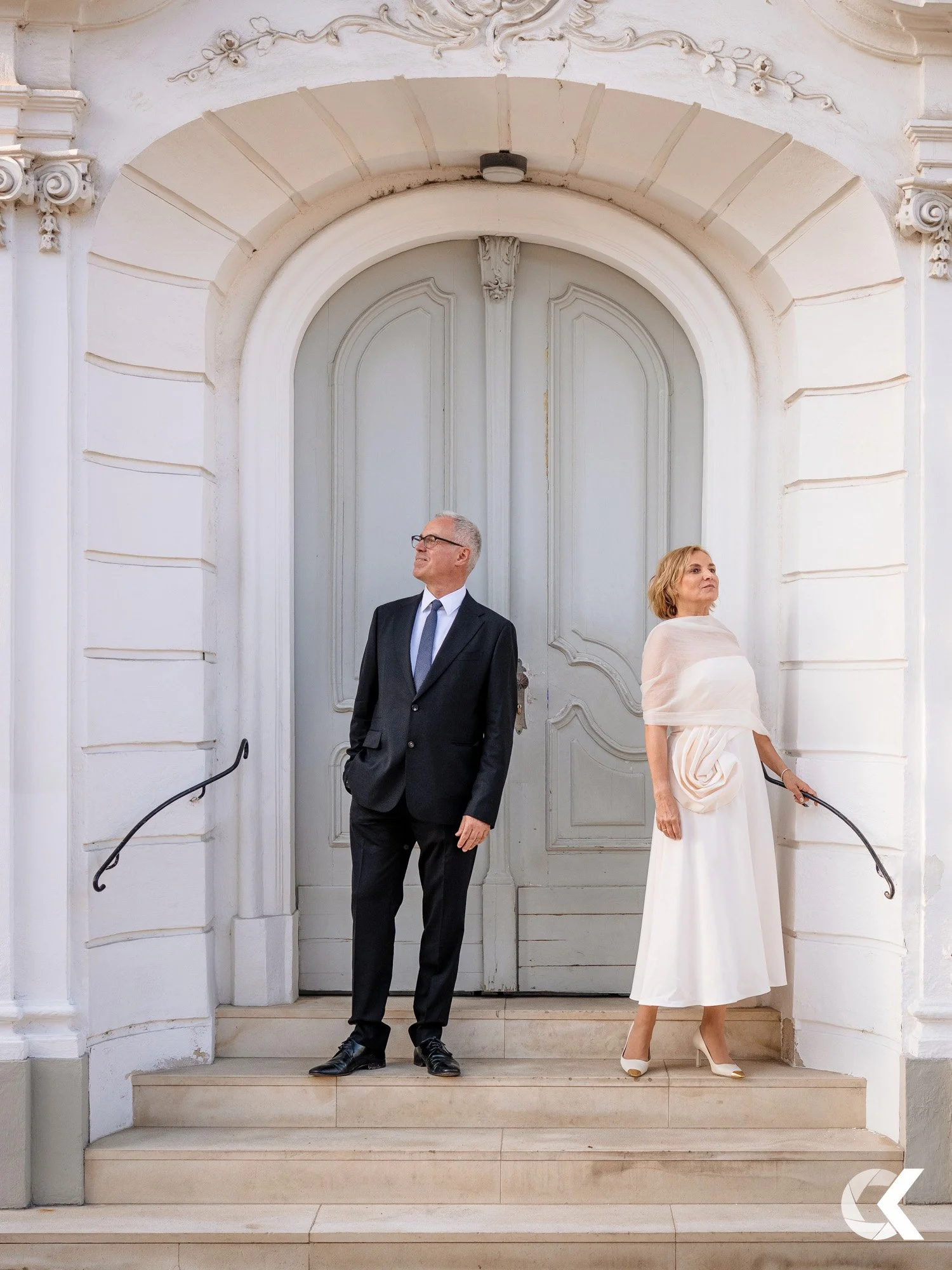 A man in a black suit and a woman in a white dress standing on steps in front of a large gray door with ornate architectural details.