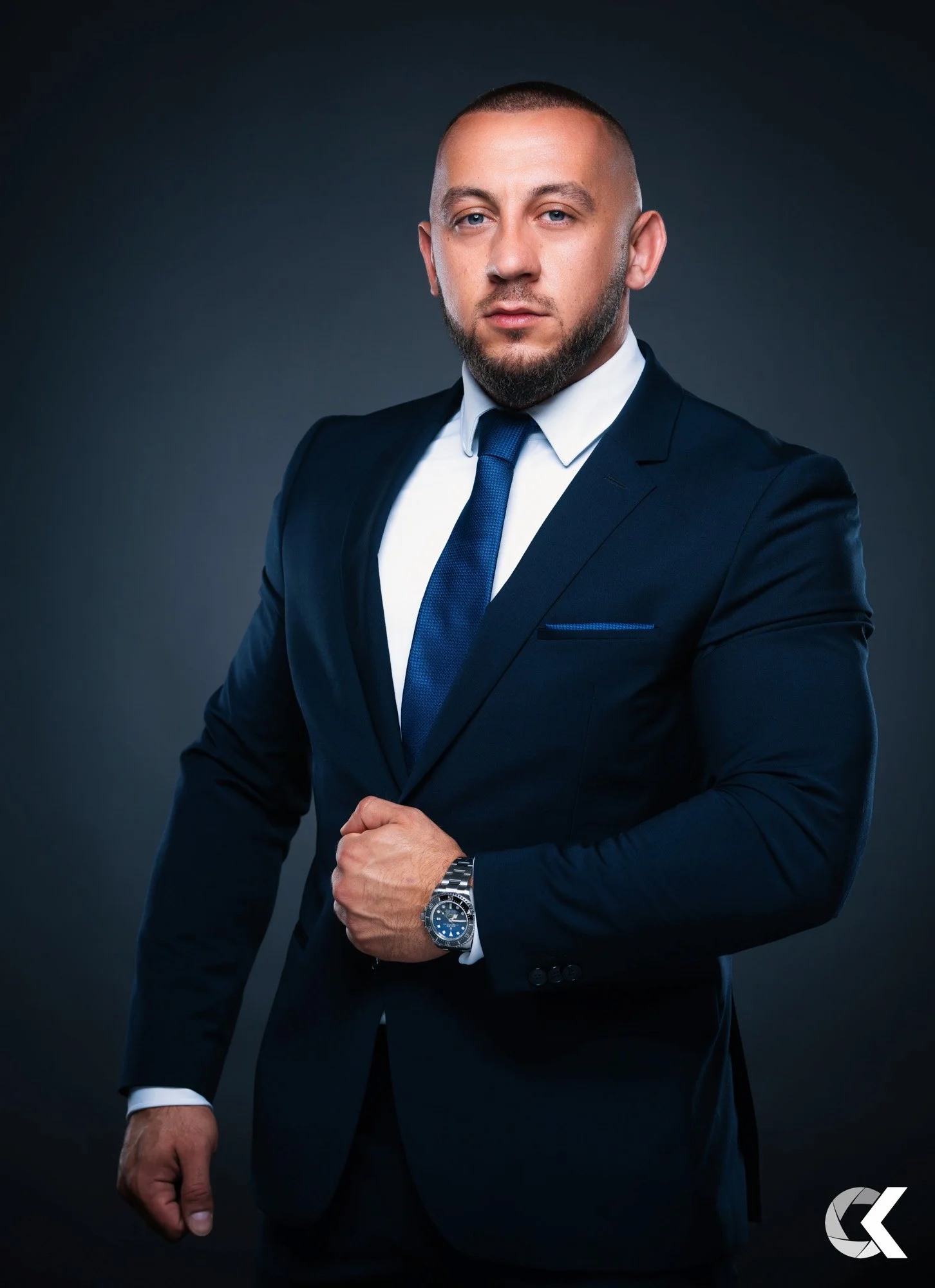 A man in a dark navy suit, white shirt, and matching tie, wearing a silver watch, standing against a dark gray background with a confident expression.