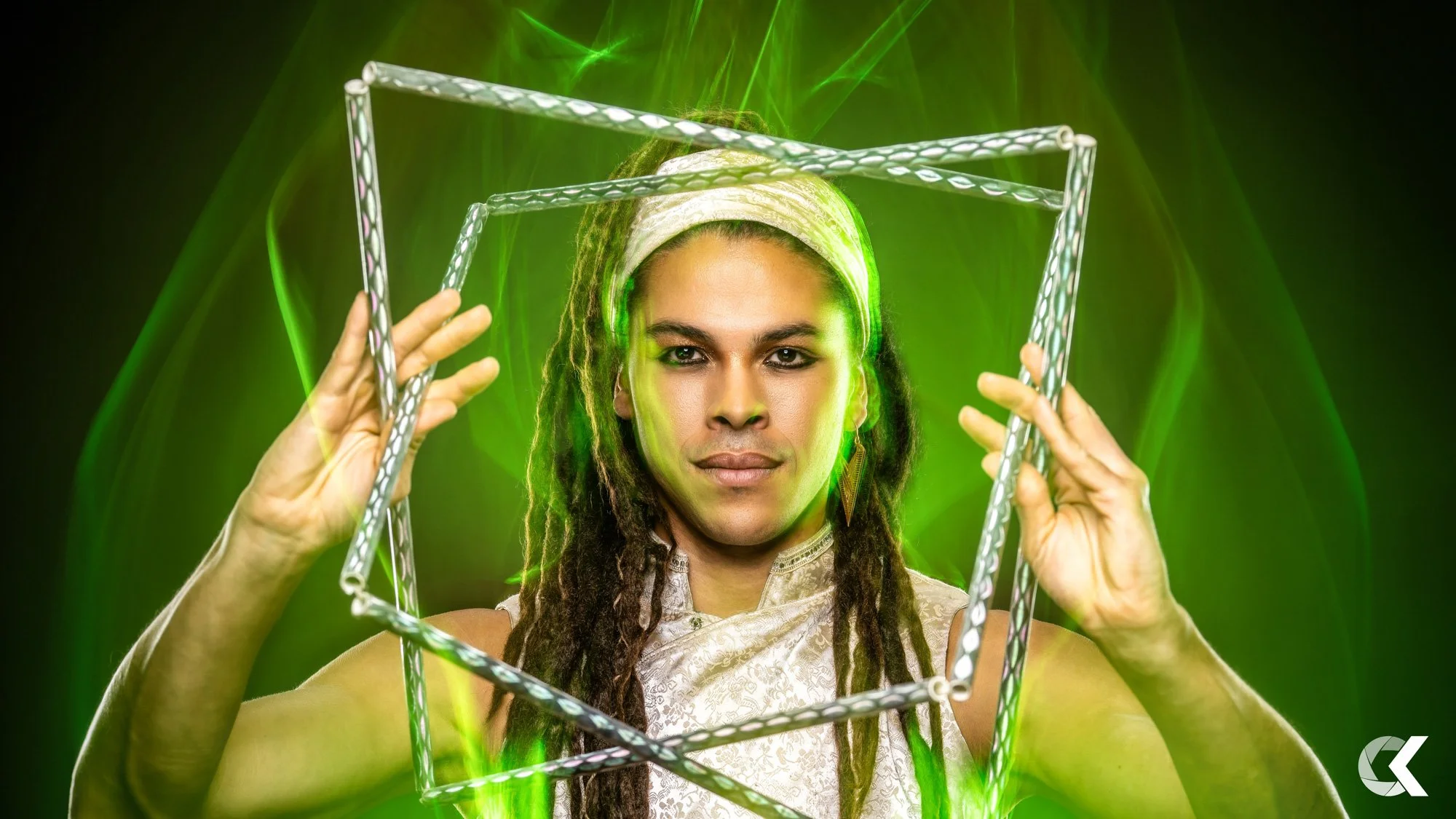 A person with long dreadlocks wearing a white headband and top, holding a metallic cube frame, with green laser lights and green lighting effects in the background.