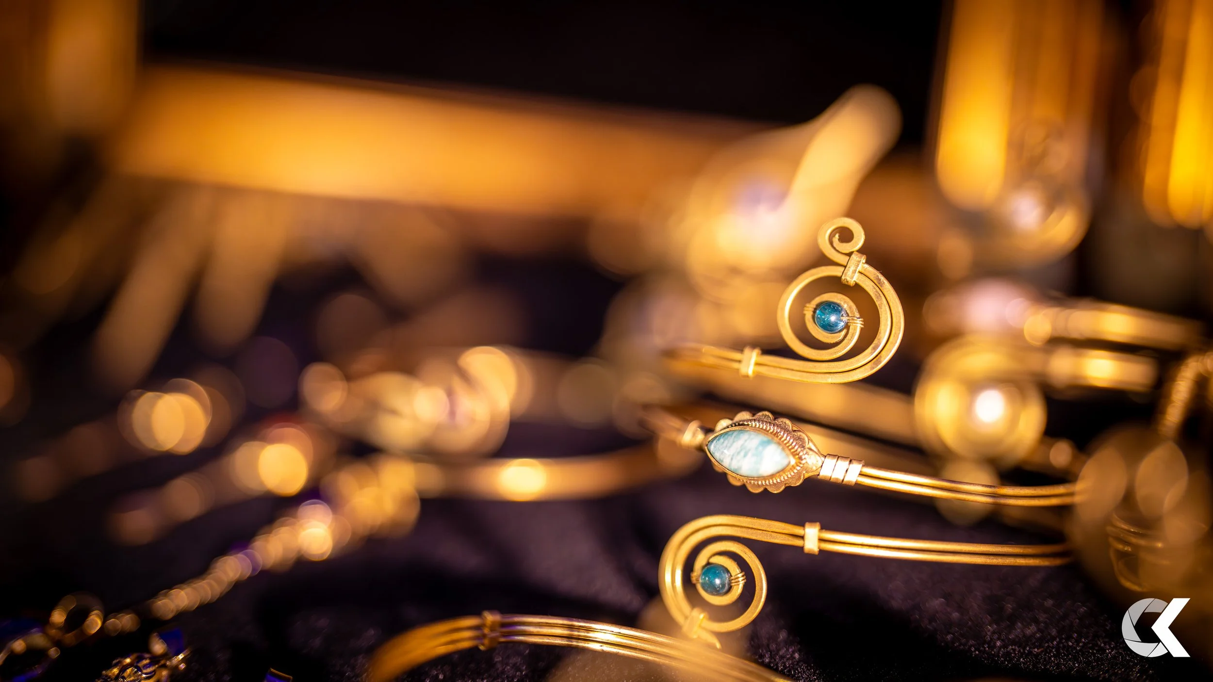 Close-up of gold jewelry pieces, including earrings and bracelets, with one featuring a blue gemstone and another with a mother-of-pearl inlay, displayed on a dark fabric.