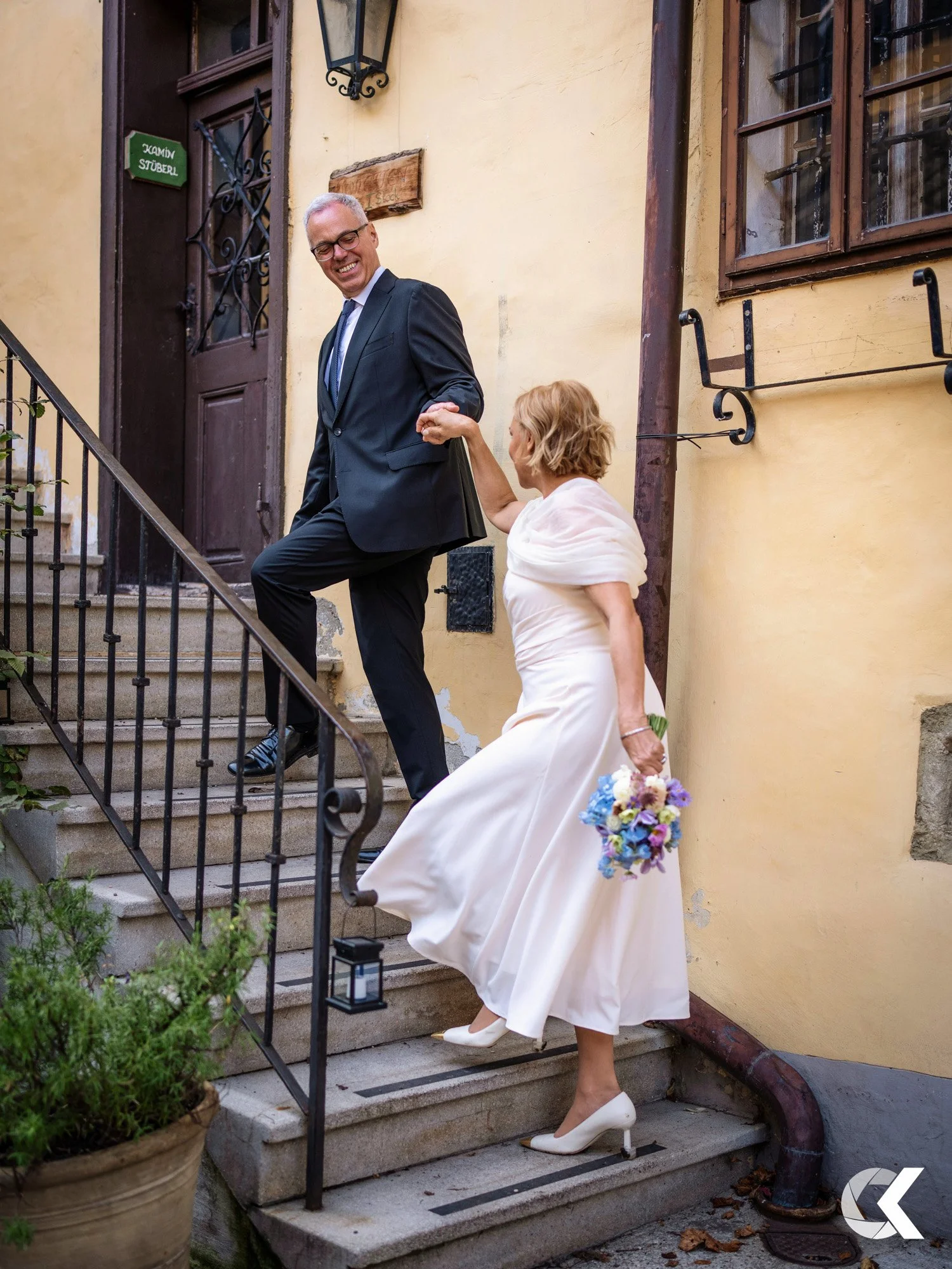 A woman in a white dress and heels is helping a man in a black suit down a set of outdoor stairs. The woman is holding a bouquet of flowers and the man is smiling.