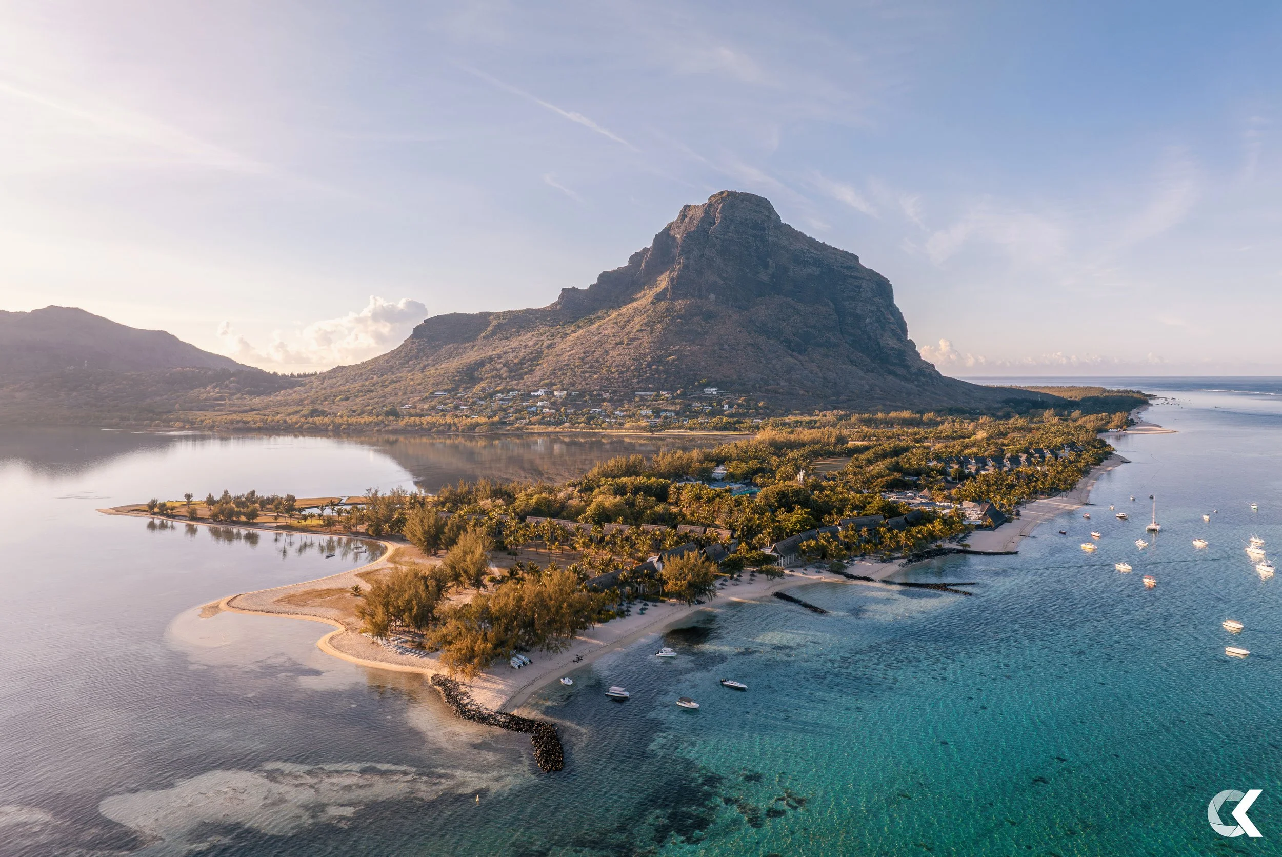 Aerial view of a coastal island with a prominent mountain, surrounded by a body of water with boats anchored near the shore, green trees, and a small beach area.