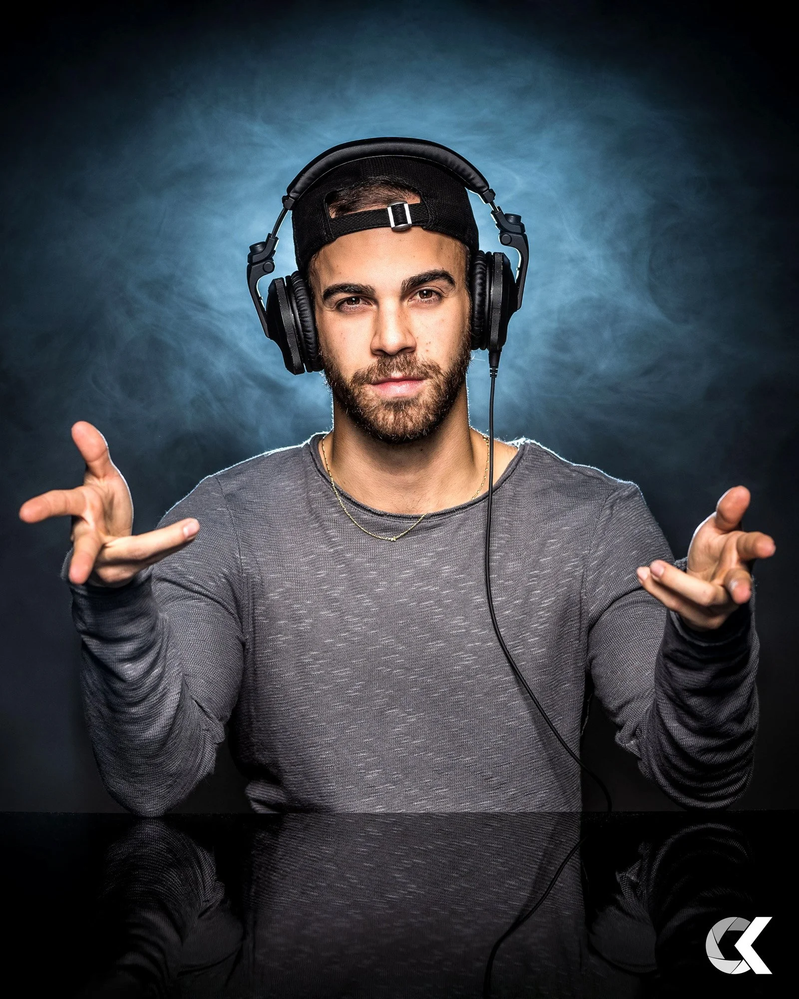 A young man with a beard wears headphones, a backwards cap, and a gray long sleeve shirt. He gestures with both hands towards the camera, standing against a textured dark background with a slight blue highlight and a reflective surface below him.