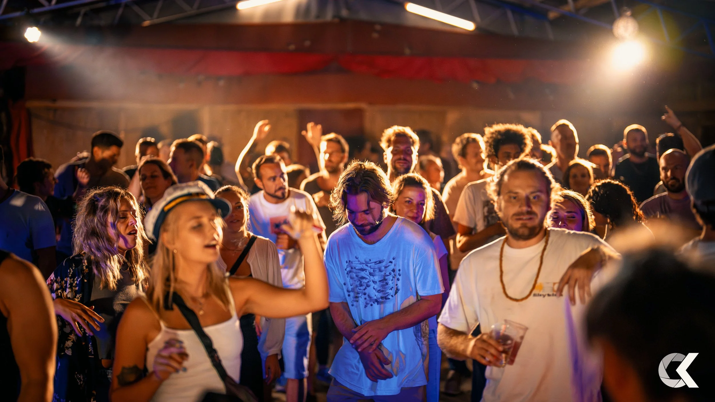A crowd of people dancing and enjoying music at an indoor music event with colorful lighting and a smoky atmosphere.