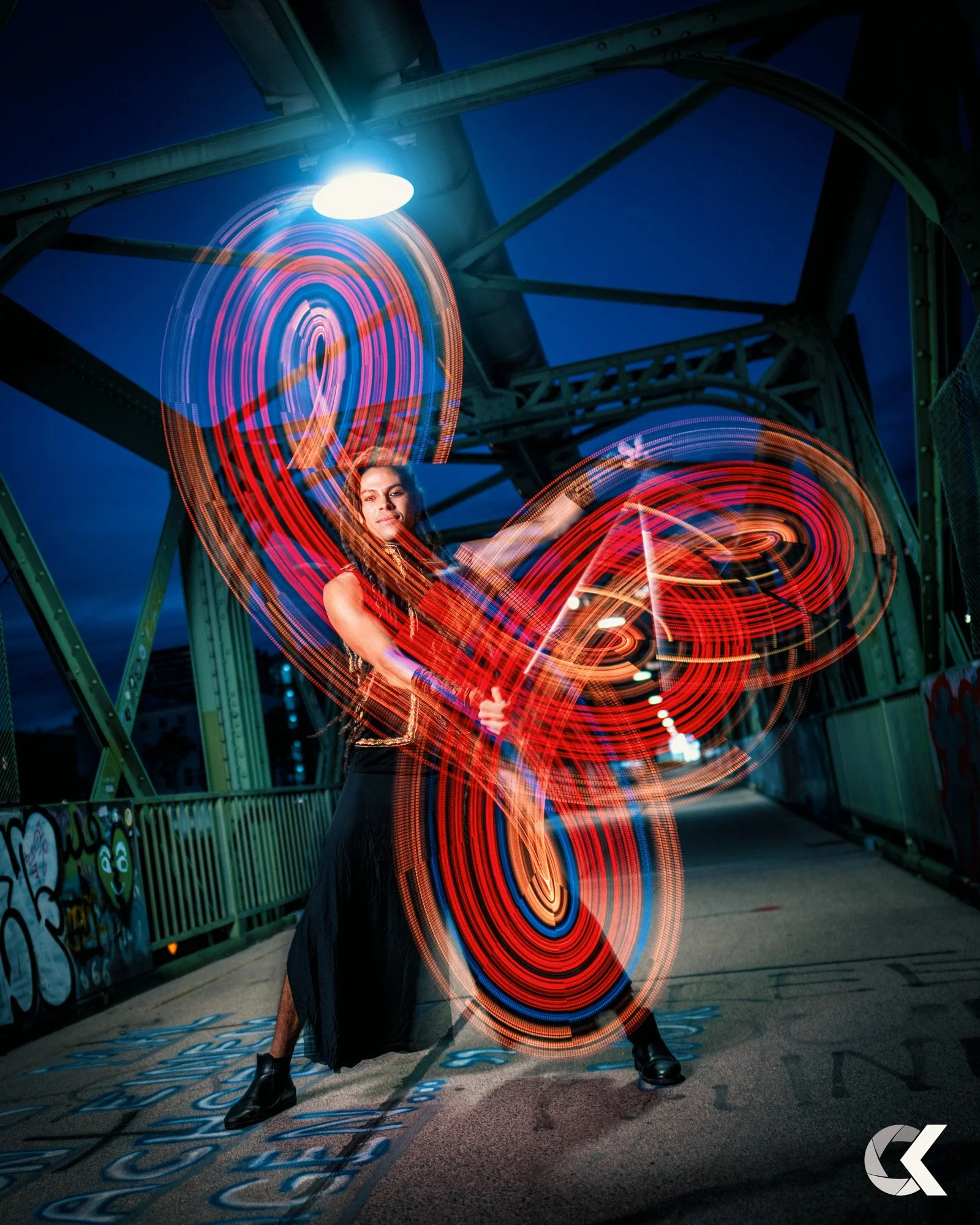 Frau auf Brücke bei Nacht, die mit Lichtmaltechnik bunte Lichterkreise und Linien zeichnet.