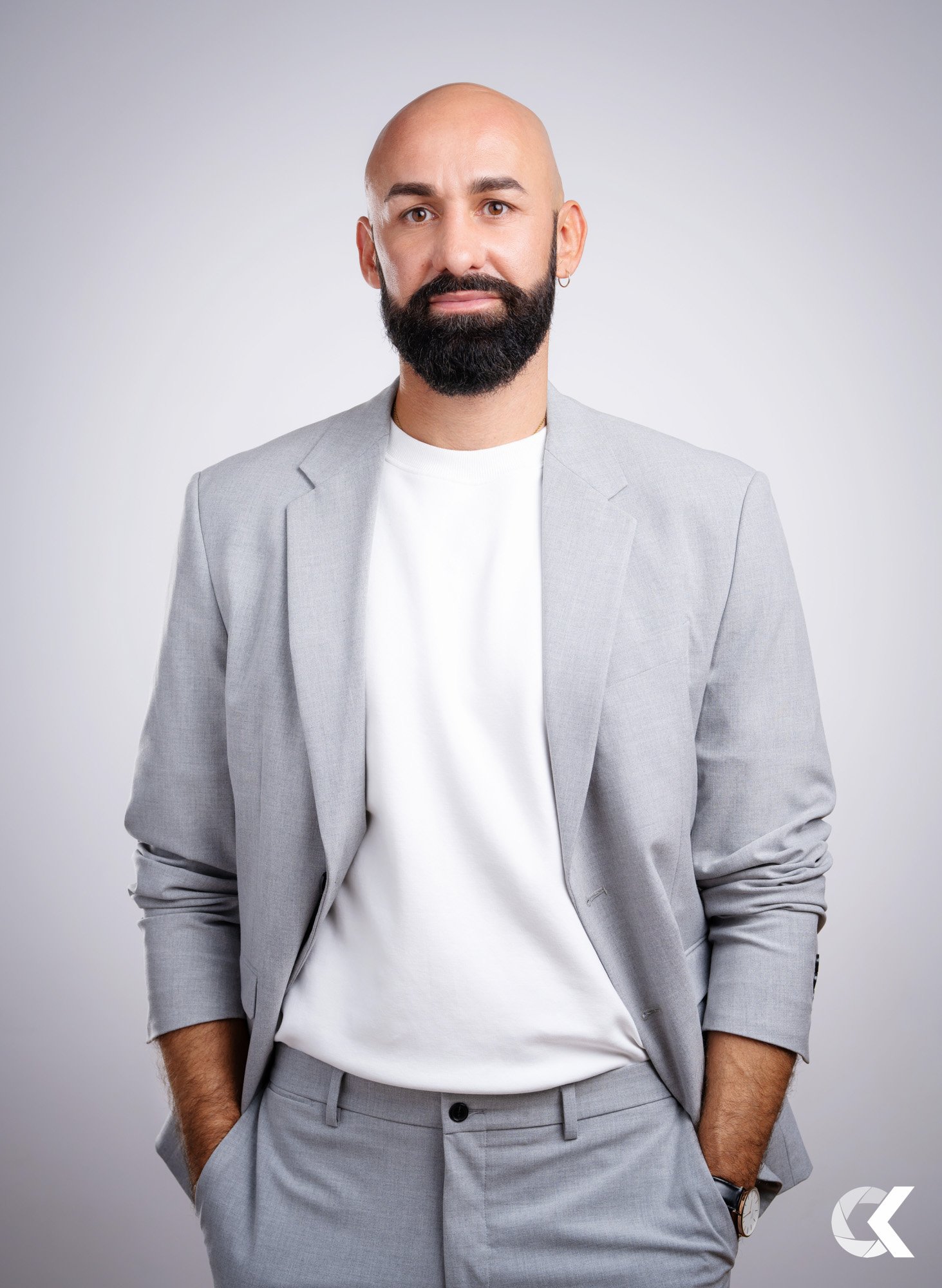 A man with a bald head, dark beard, and earring, wearing a light gray suit with a white shirt, stands against a plain gray background with his hands in his pockets.