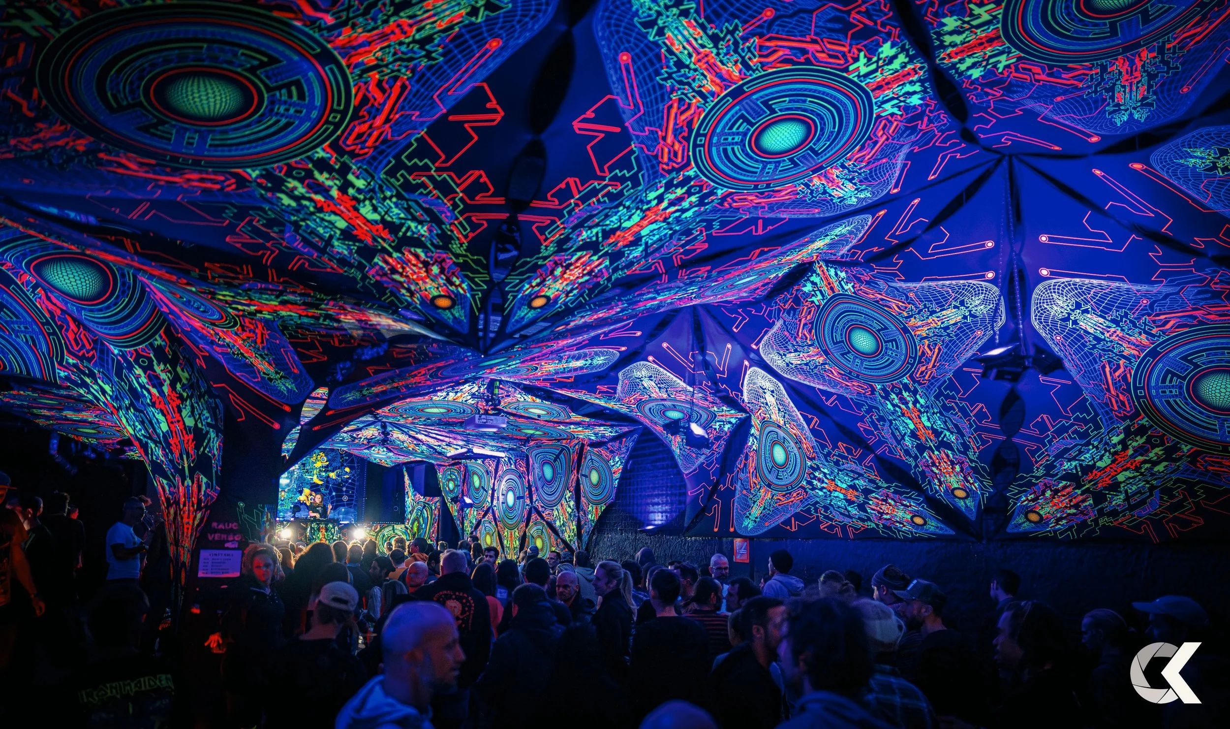 Crowd of people at a night event with neon-colored, abstract, circuit-like patterns on the ceiling and walls.