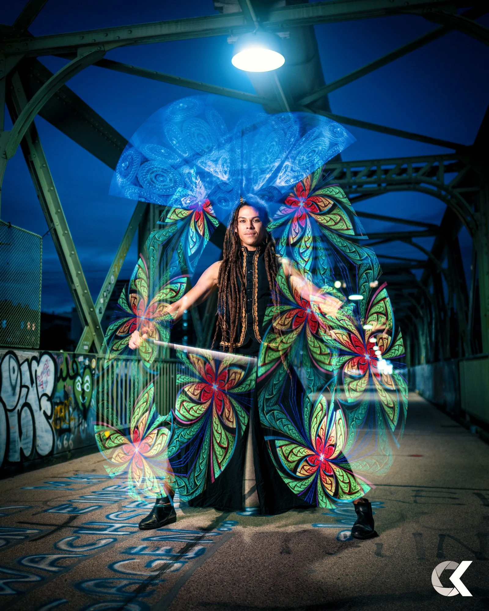 Eine Person mit langen dreadlocks trägt ein leuchtendes Outfit mit bunten, fluoreszierenden Butterfly-Mustern unter einer Brücke in der Nacht.