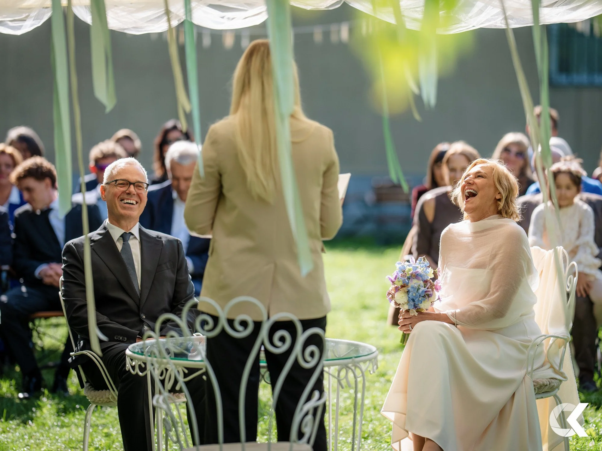 A wedding ceremony outdoors with a bride and groom seated, laughing and smiling, while a woman officiates. Guests are seated in the background.