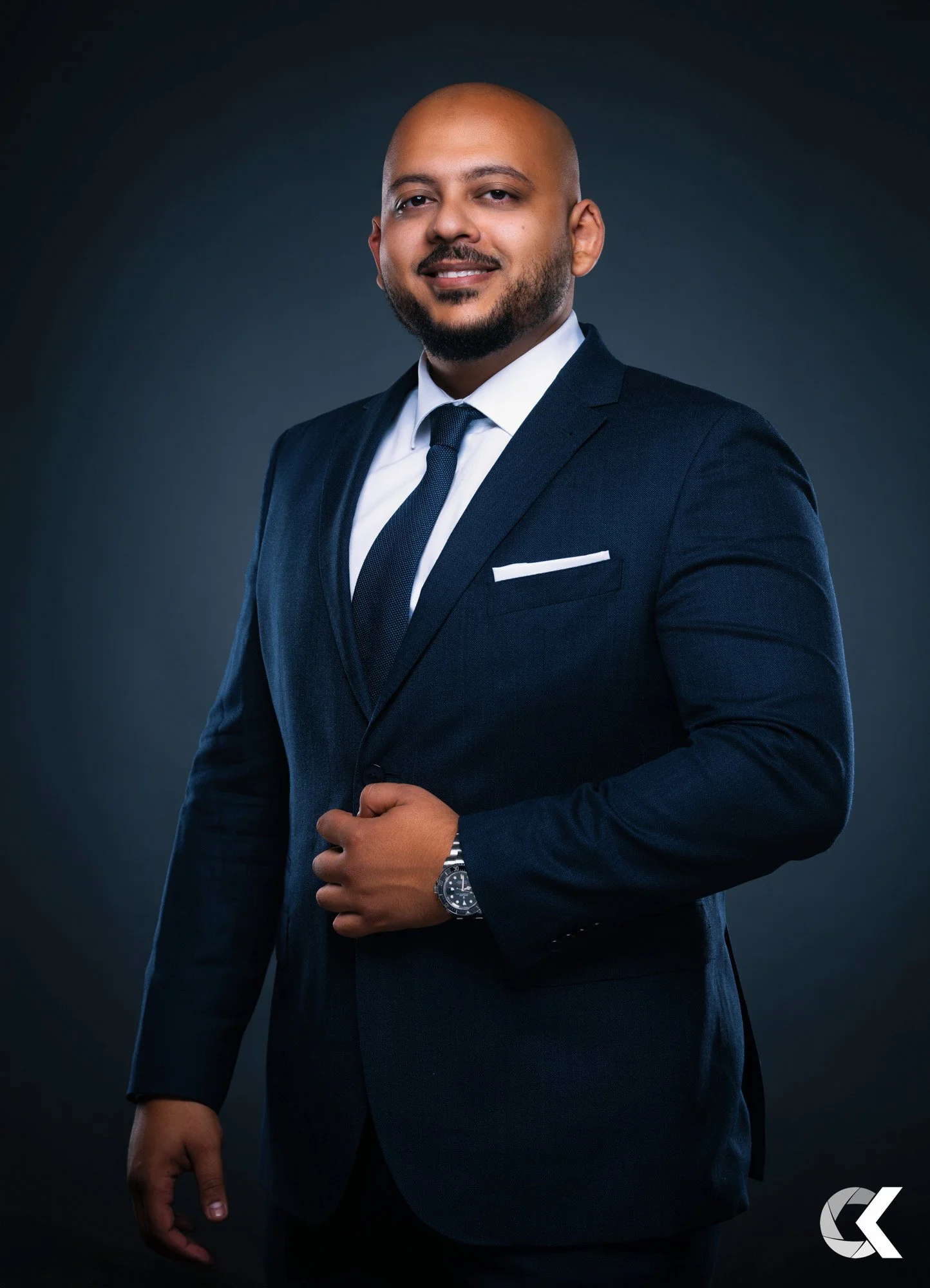 A portrait of a man in a navy blue suit, white shirt, and dark tie, standing against a dark background. He has a beard, is smiling slightly, and is wearing a wristwatch.