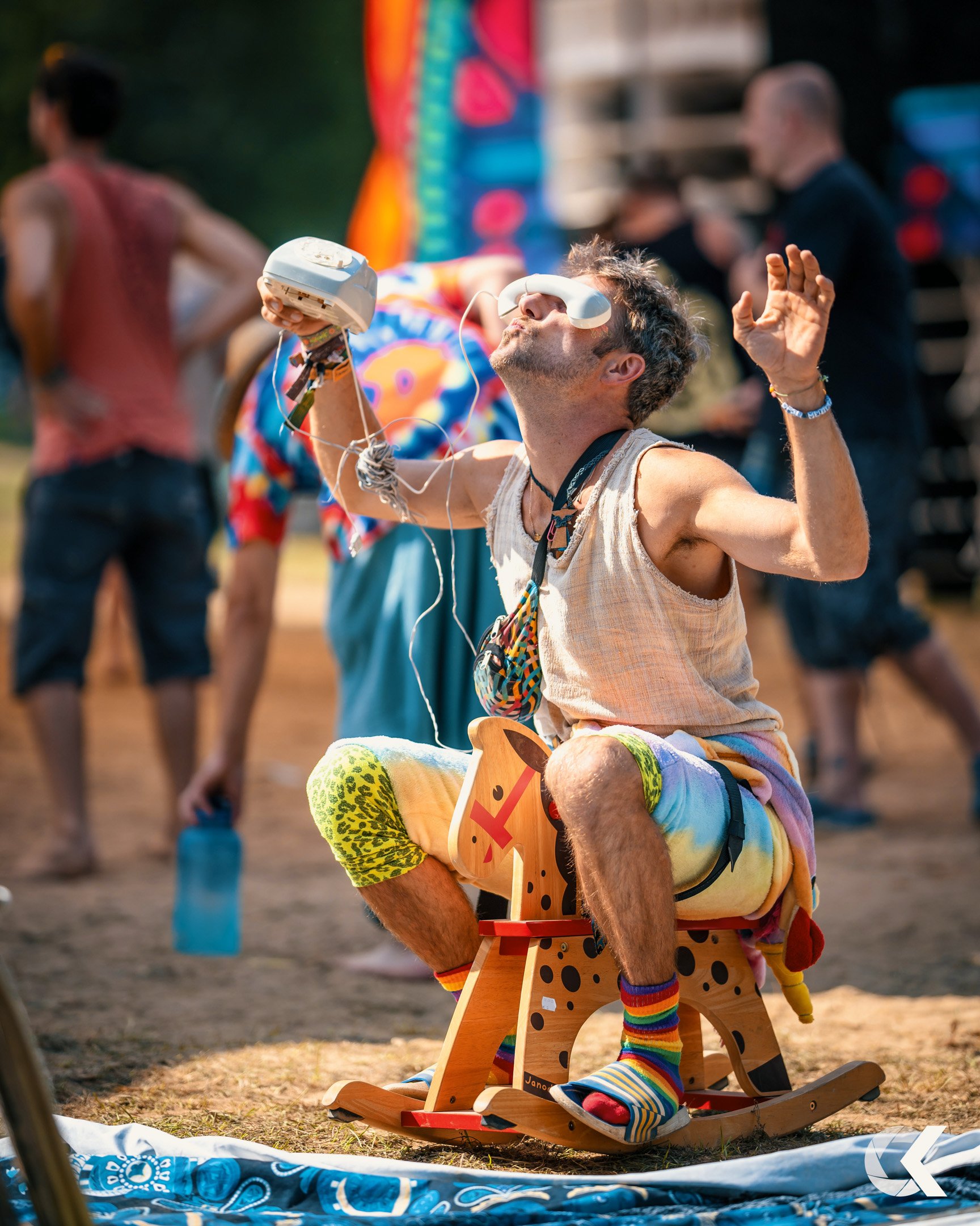 Man sitting on a wooden rocking horse, wearing colorful socks and shorts, holding a retro telephone receiver with tangled wires, in a lively outdoor festival setting.