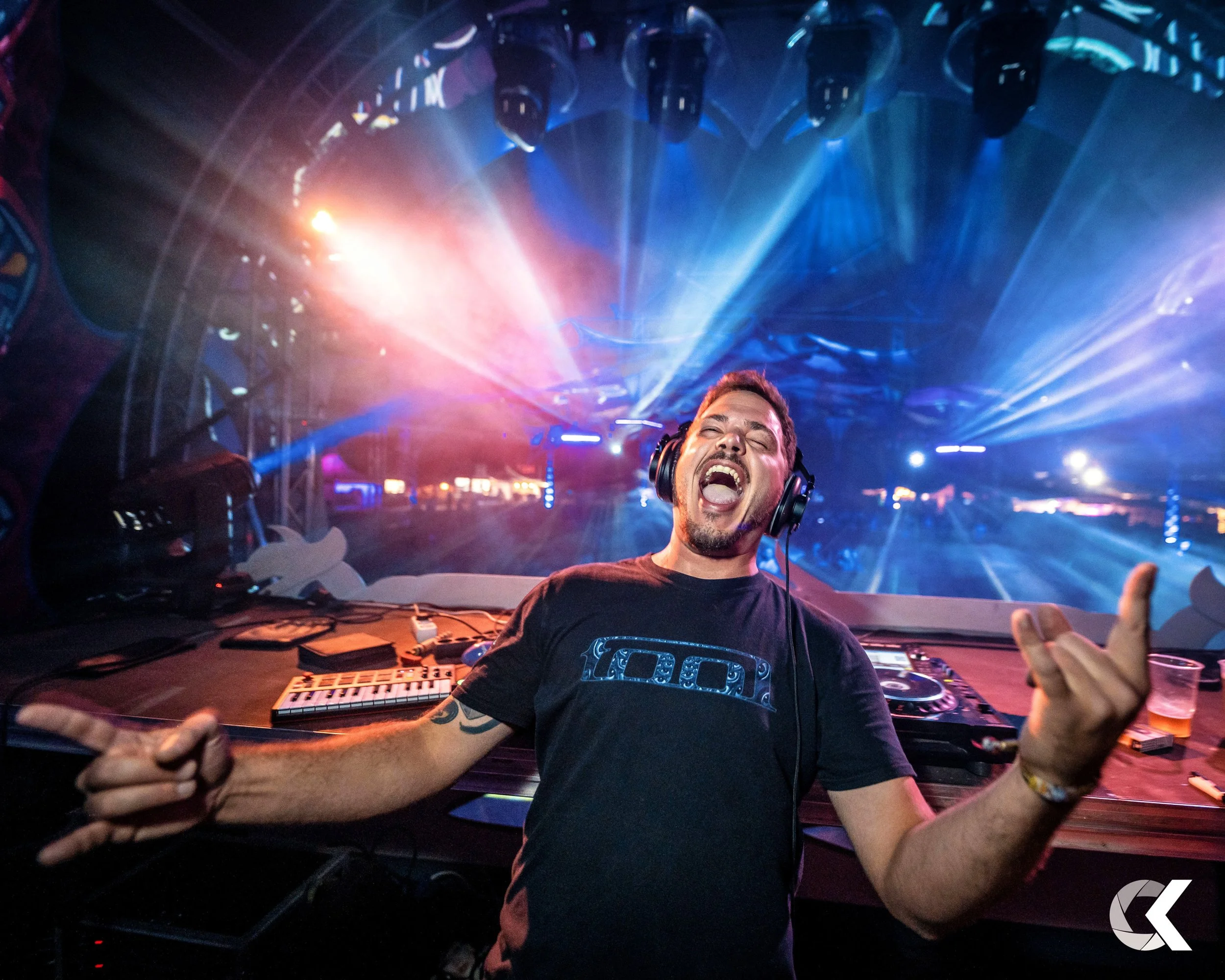 A DJ in a black t-shirt with a design on it, wearing headphones, is singing and enjoying himself in front of a DJ setup with colorful stage lights and beams in the background.