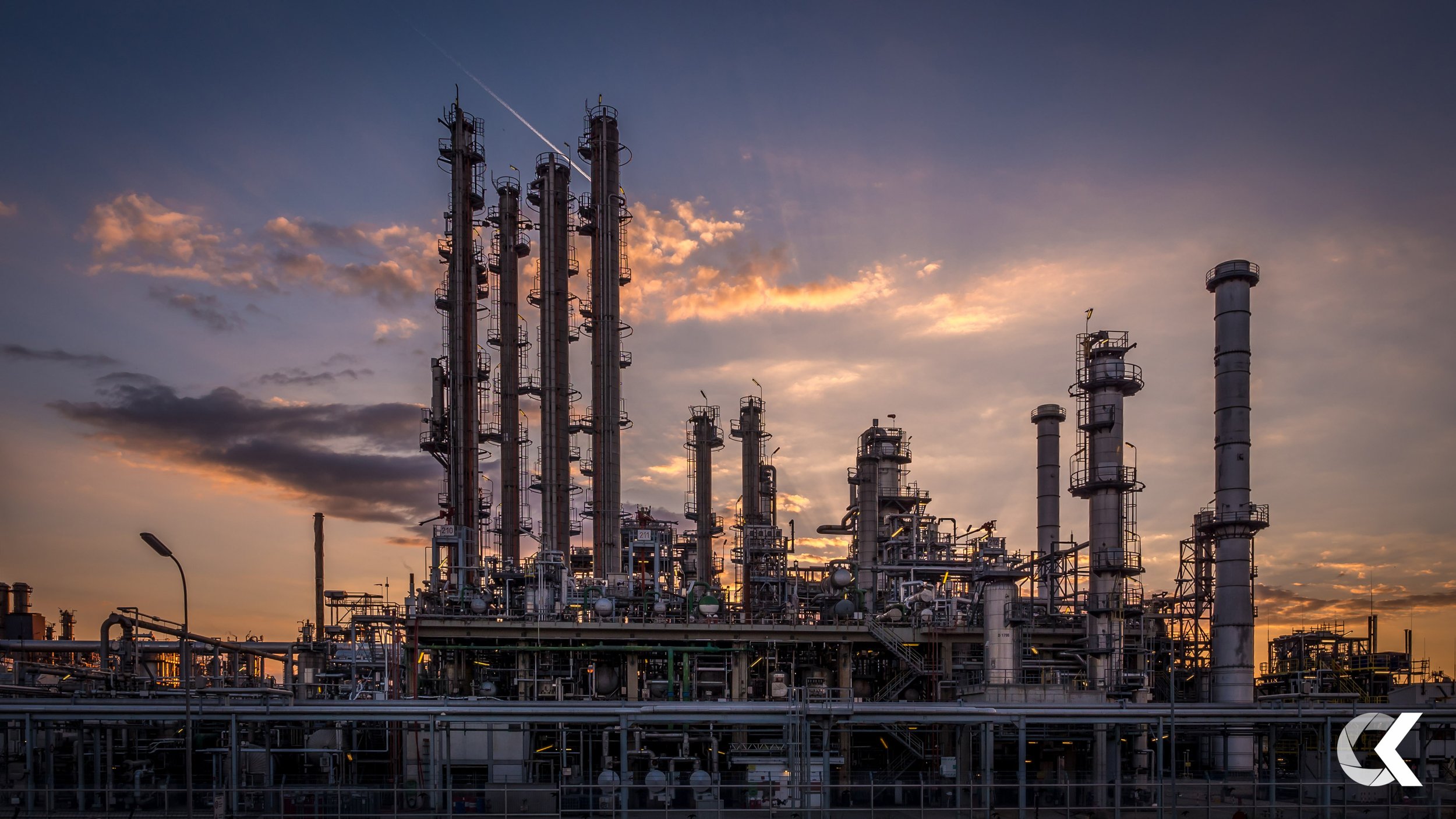 An industrial oil refinery with multiple tall towers and complex piping, set against a sunset sky with clouds.
