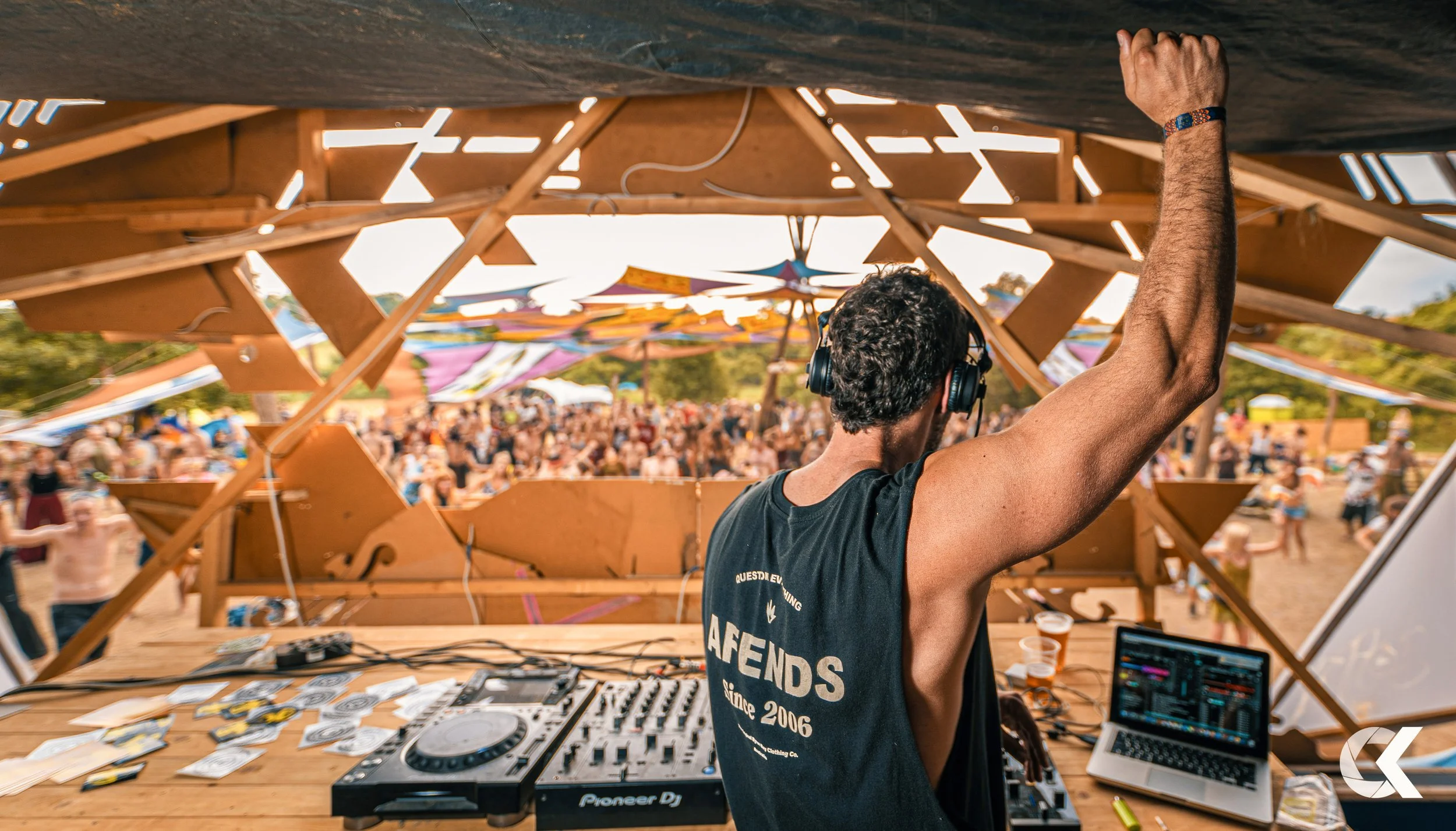 DJ performing at an outdoor music festival with a large crowd in the background. The DJ is wearing headphones and has one arm raised. There is DJ equipment and a laptop on the table, and the stage is made of wood with colorful fabric decorations over