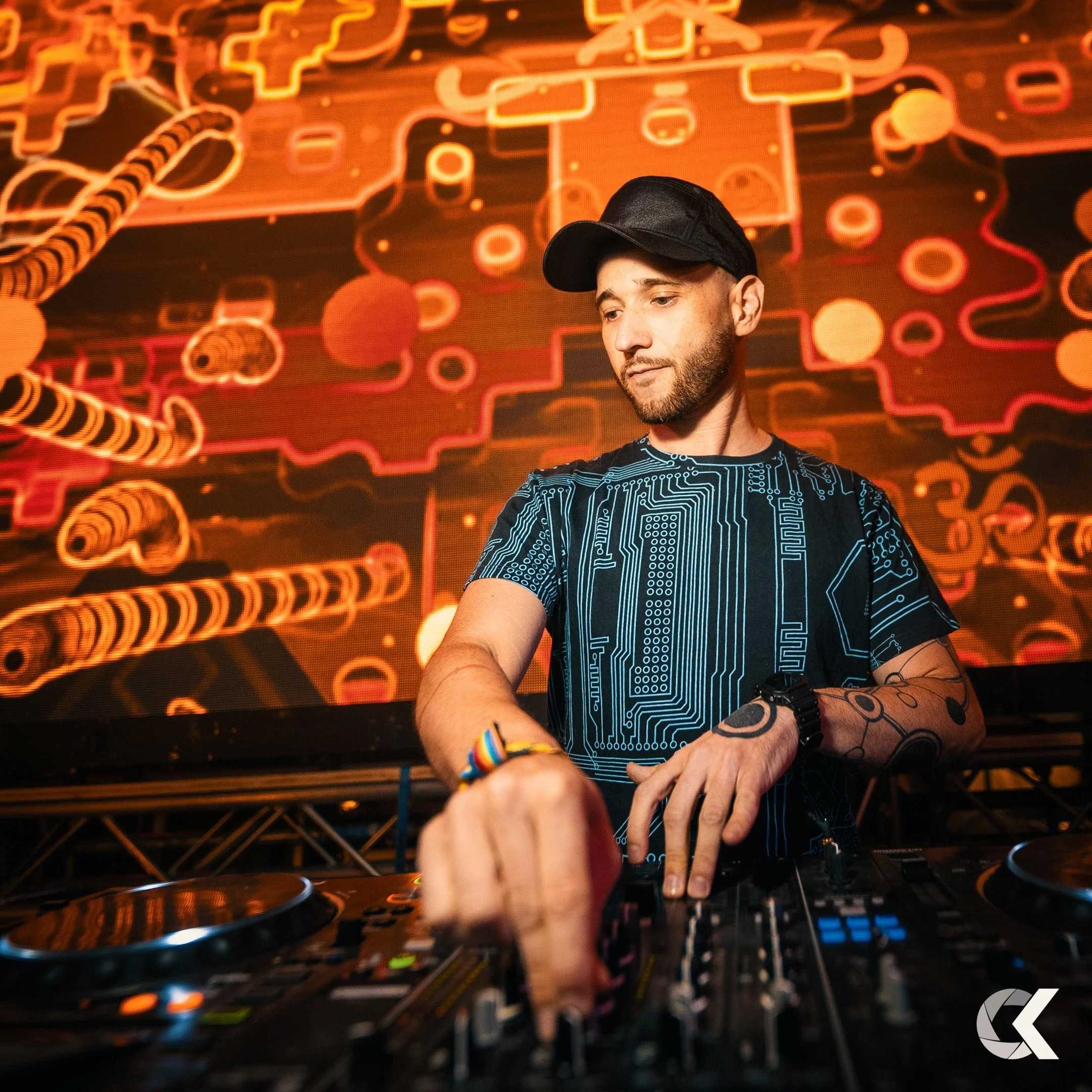 A DJ wearing a black cap and a blue circuit board patterned shirt mixes music at a nightclub with a vibrant digital abstract background.