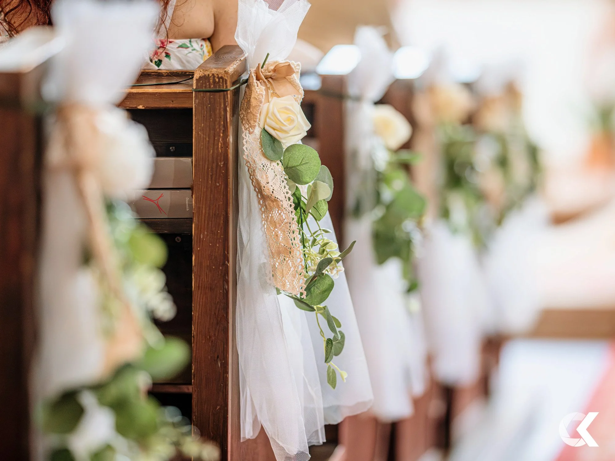 Wooden pew decorated with white tulle, lace, and greenery, including white roses, at a wedding ceremony.