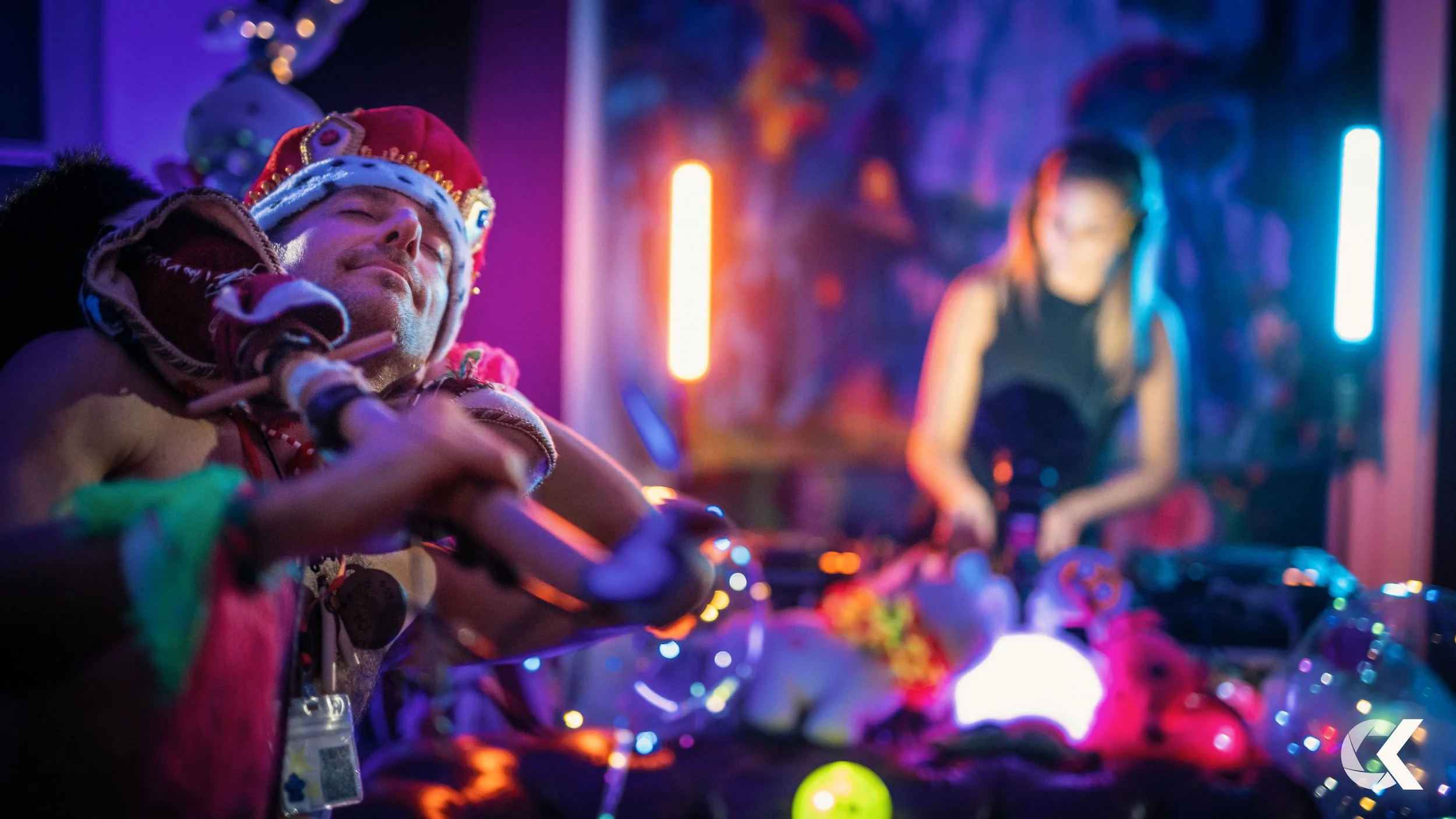 A person dressed as a king with a red crown and royal robe, smiling with eyes closed, in a colorful, neon-lit party environment, with a DJ woman in the background.