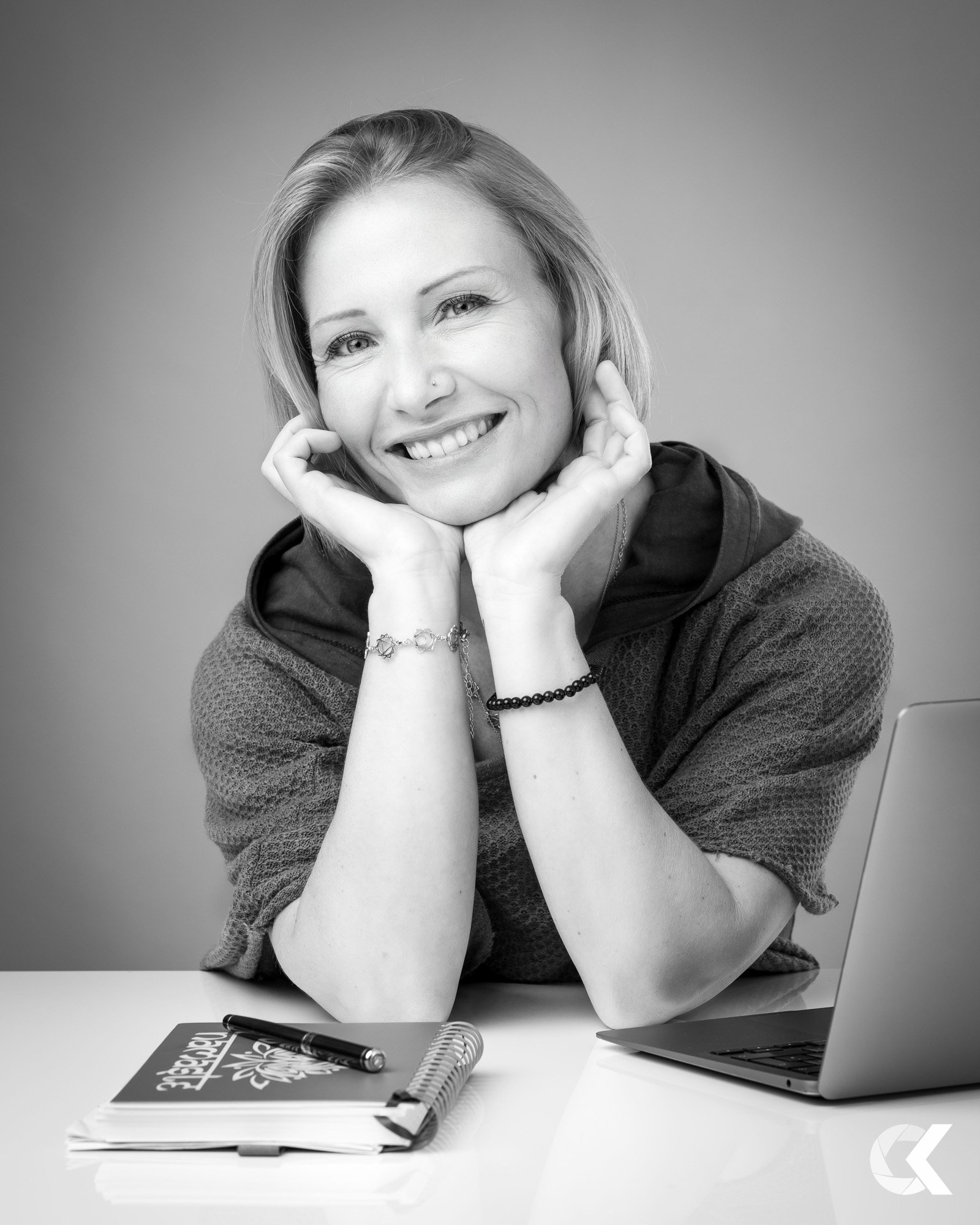 A smiling woman with short hair resting her chin on her hands, sitting at a desk with a laptop, notebook, and pen.