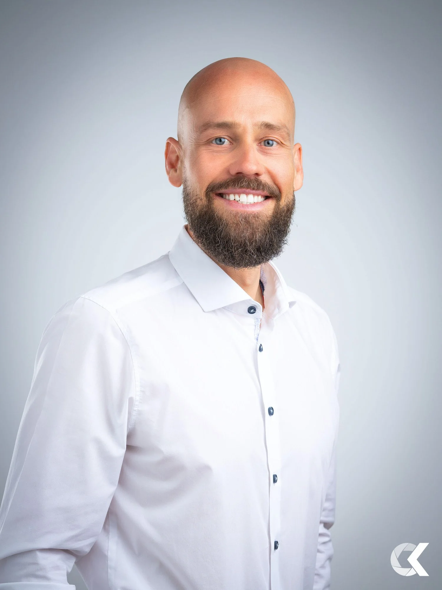 Portrait of a smiling man with a bald head and full beard, wearing a white button-up shirt against a light gray background.