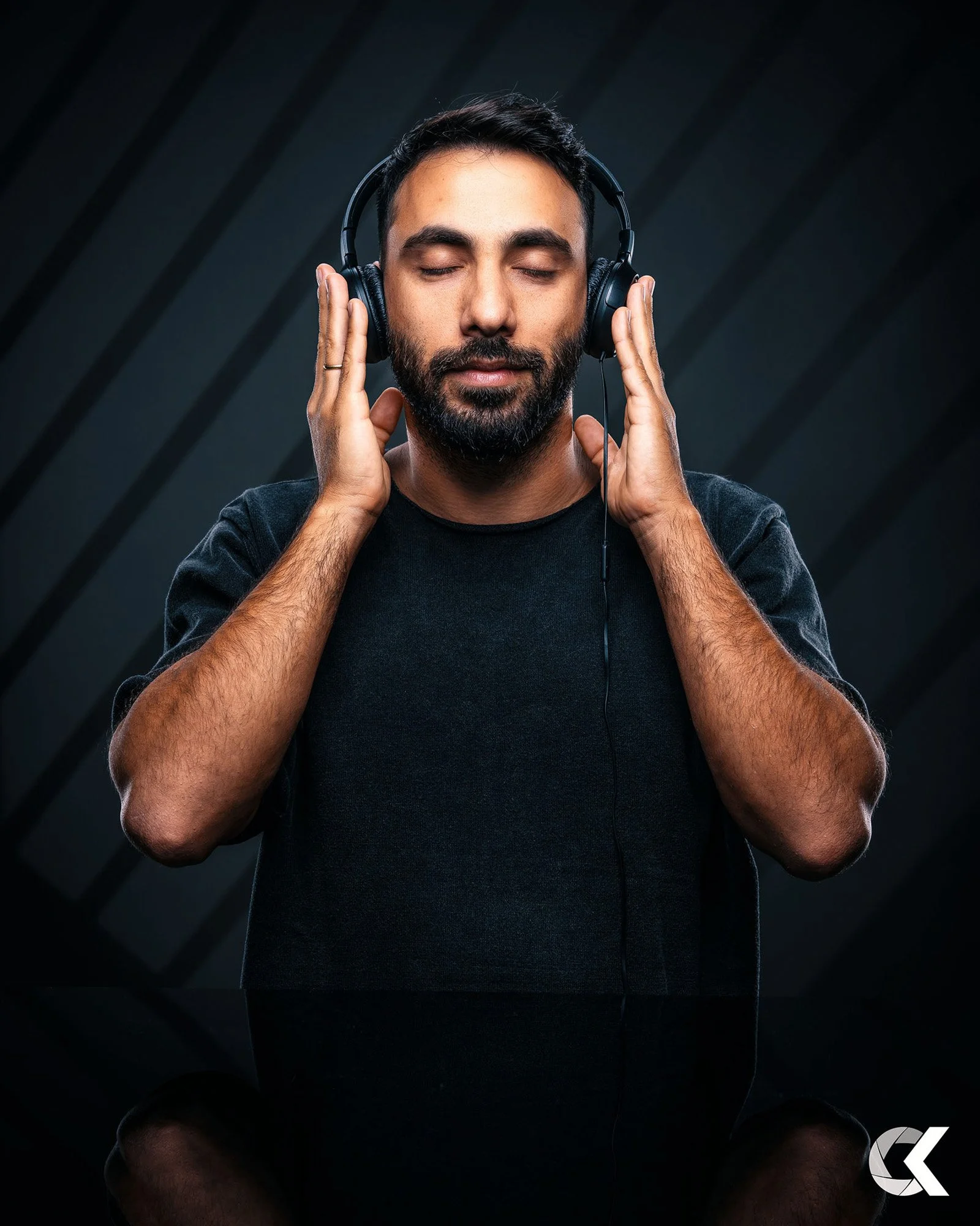 A man with a beard and dark hair wearing headphones with eyes closed, listening to music or audio in a dark studio setting.