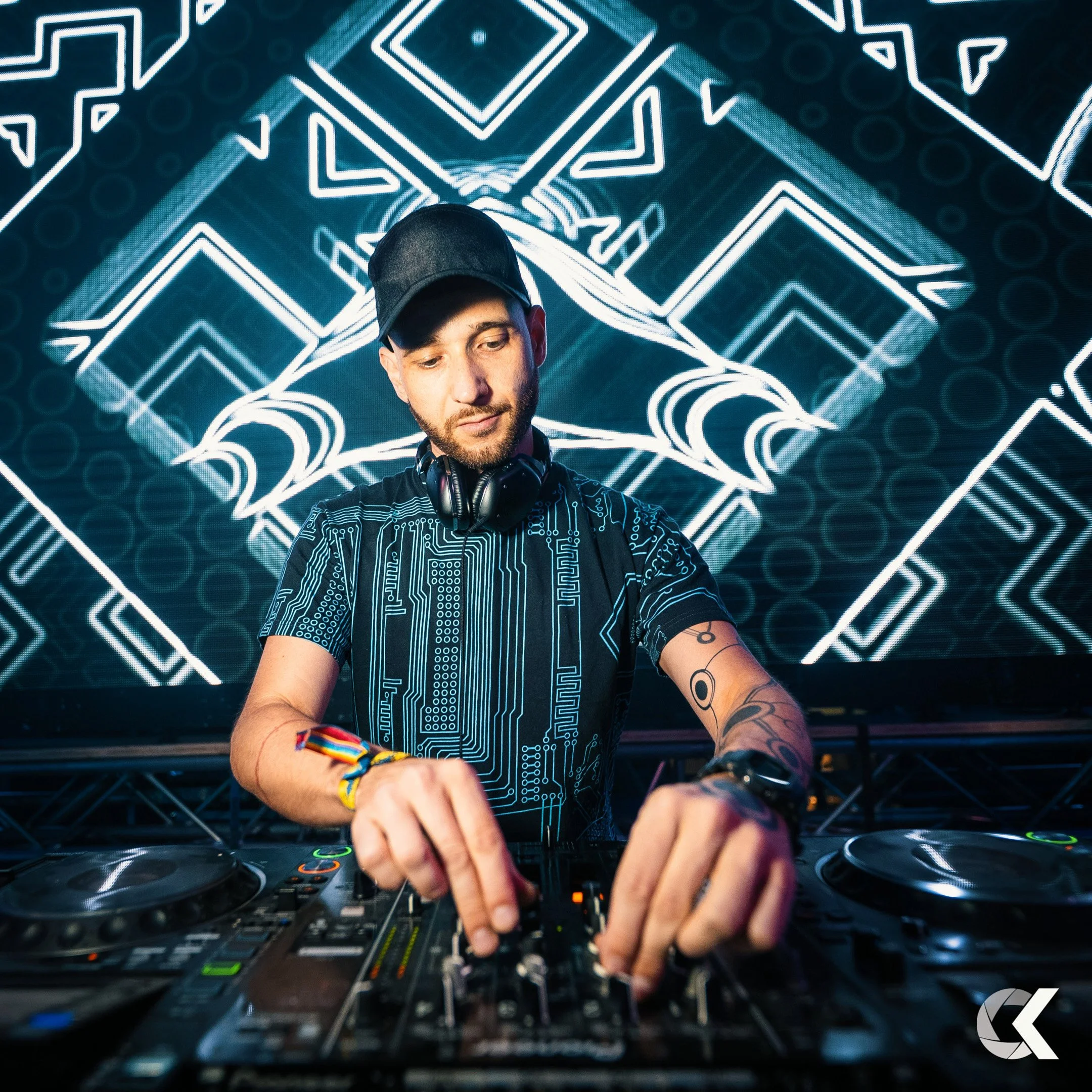 A DJ wearing a black cap, headphones around neck, and a black shirt with blue circuit board patterns, is operating DJ equipment with colorful lighting and a digital geometric pattern on a large screen behind him.