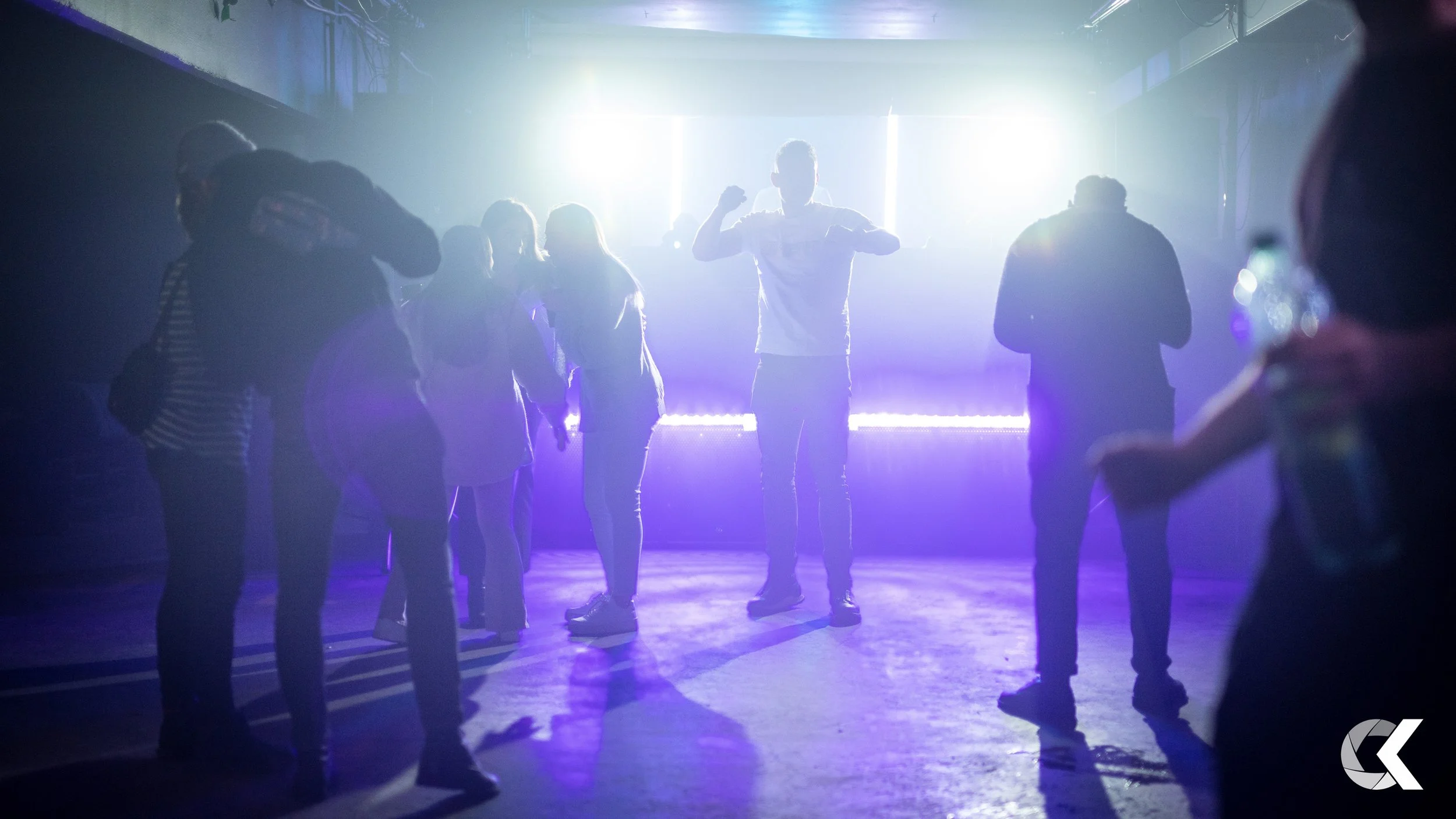 People dancing at a nightclub or concert with vibrant purple and blue lights and a bright white backlight.