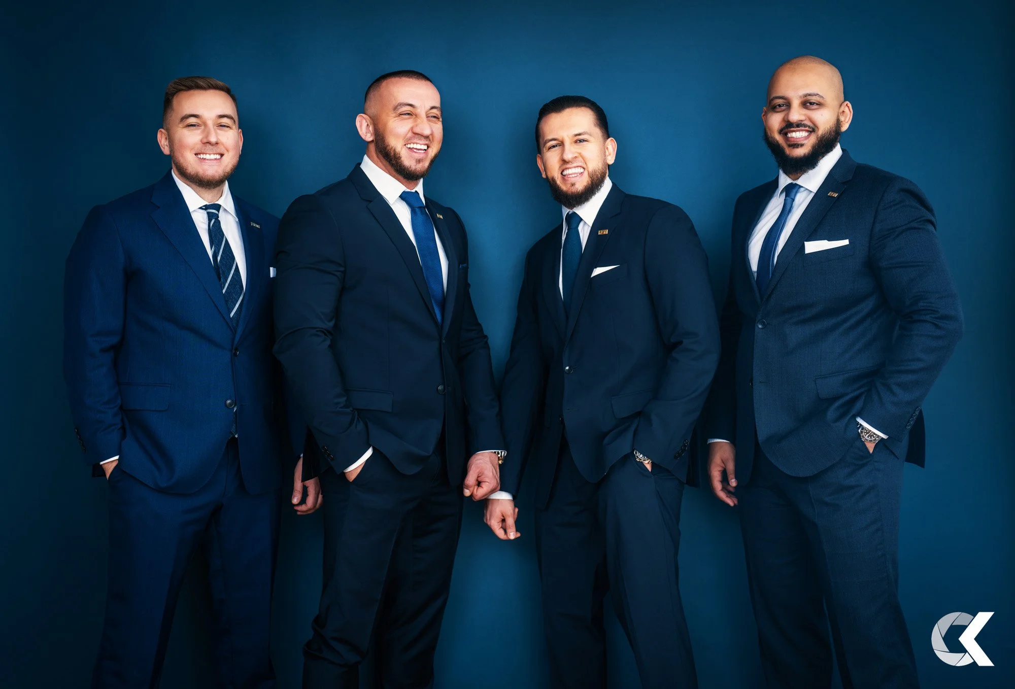Four men in dark blue suits smiling and standing against a blue background.