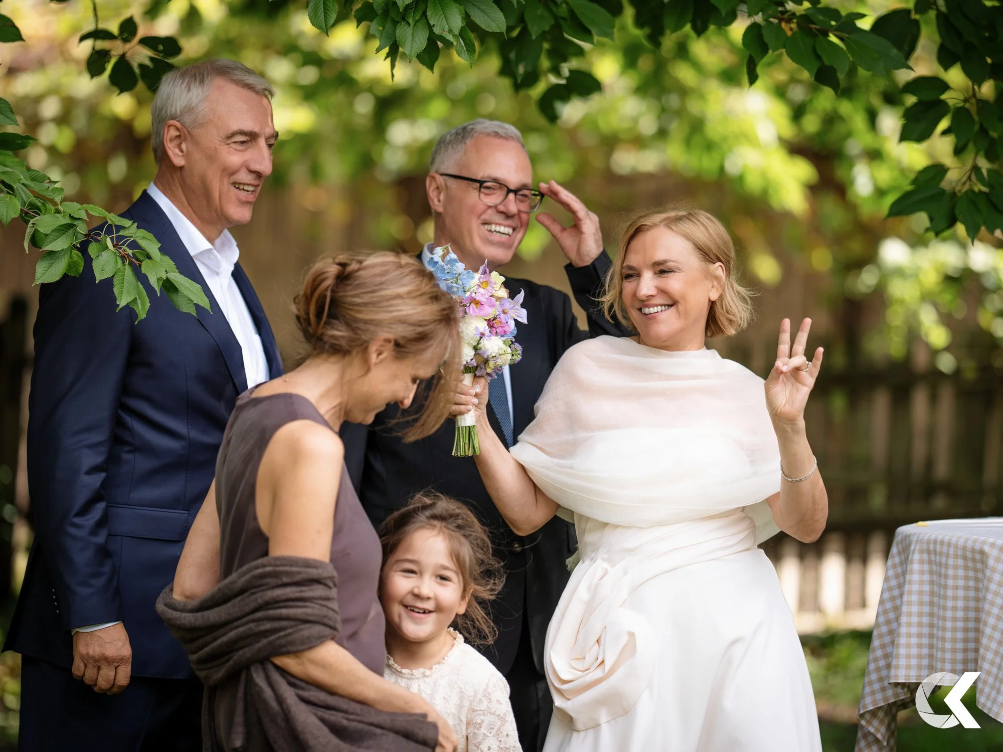 A group of five people, including two men, two women, and a young girl, smiling and enjoying an outdoor gathering with green trees overhead.
