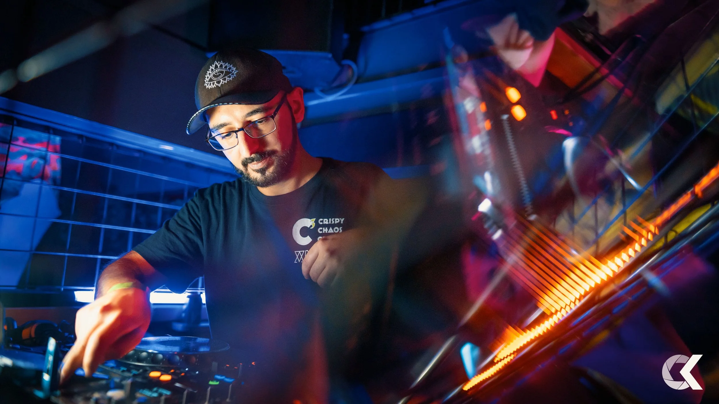 A DJ with glasses and a beard is operating DJ equipment at a nightclub, illuminated by blue and orange lights, wearing a black cap and t-shirt.