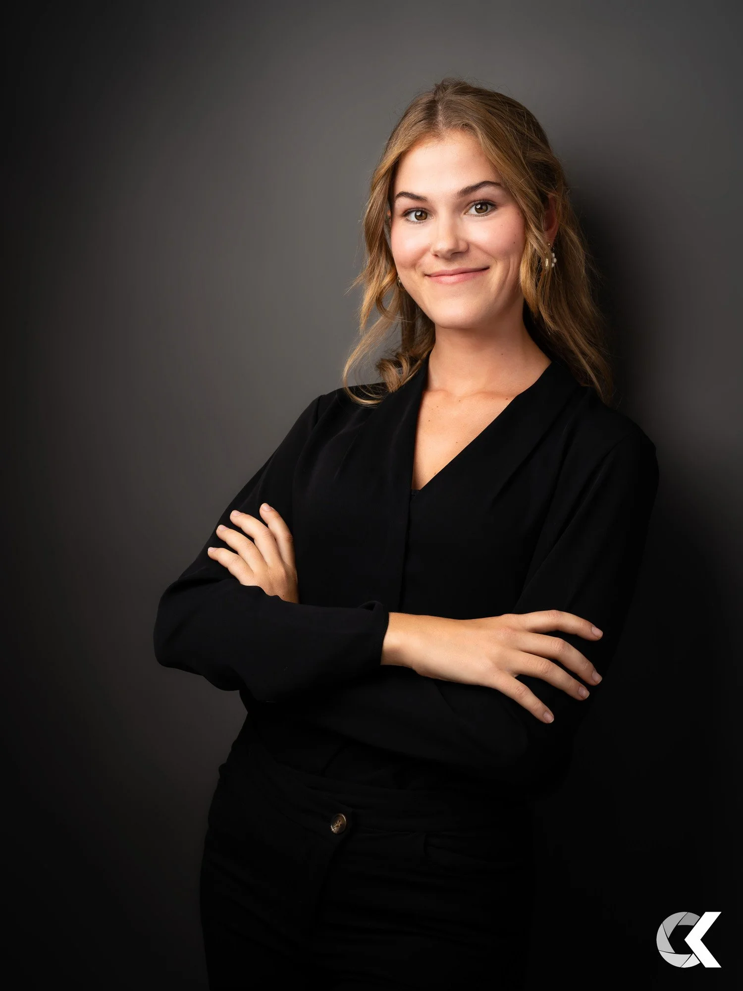 A young woman with wavy light brown hair, wearing a black long-sleeve top, smiling with arms crossed, standing against a dark grey background.