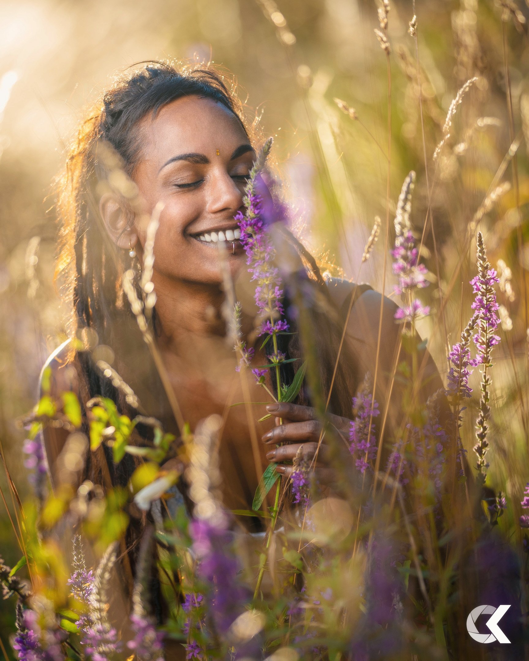 Smiling woman with long braided hair, holding purple wildflowers, in a sunlit field of tall grasses and wildflowers.