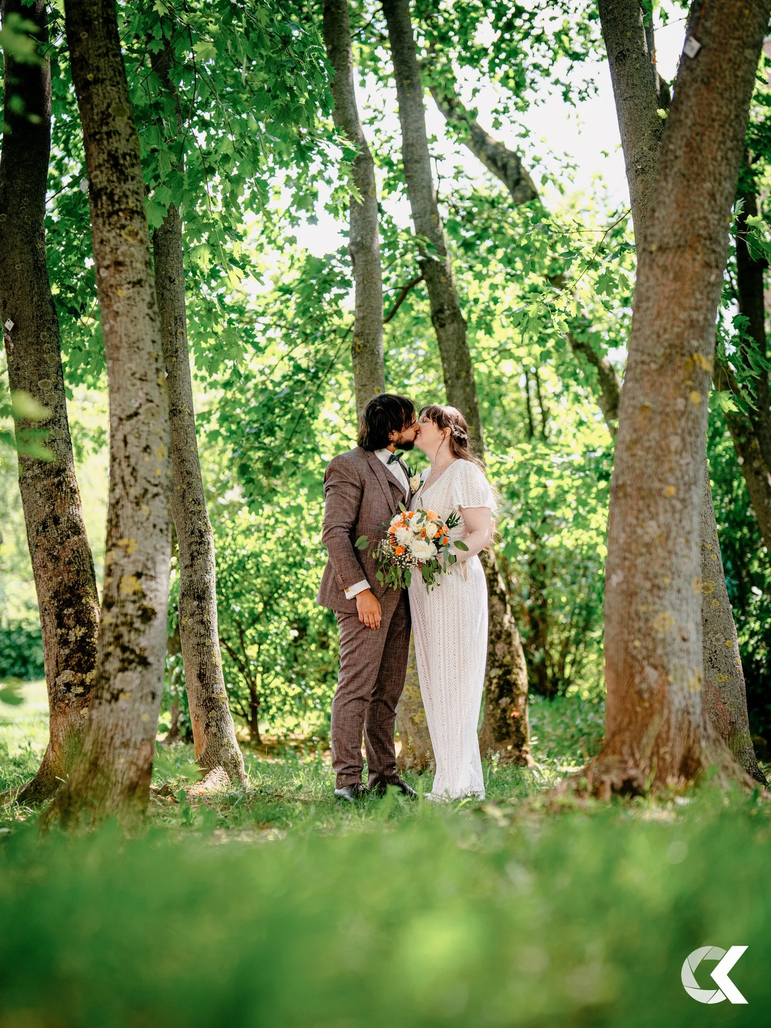 A couple in wedding attire sharing a kiss in a wooded area with green foliage and sunlight filtering through trees.