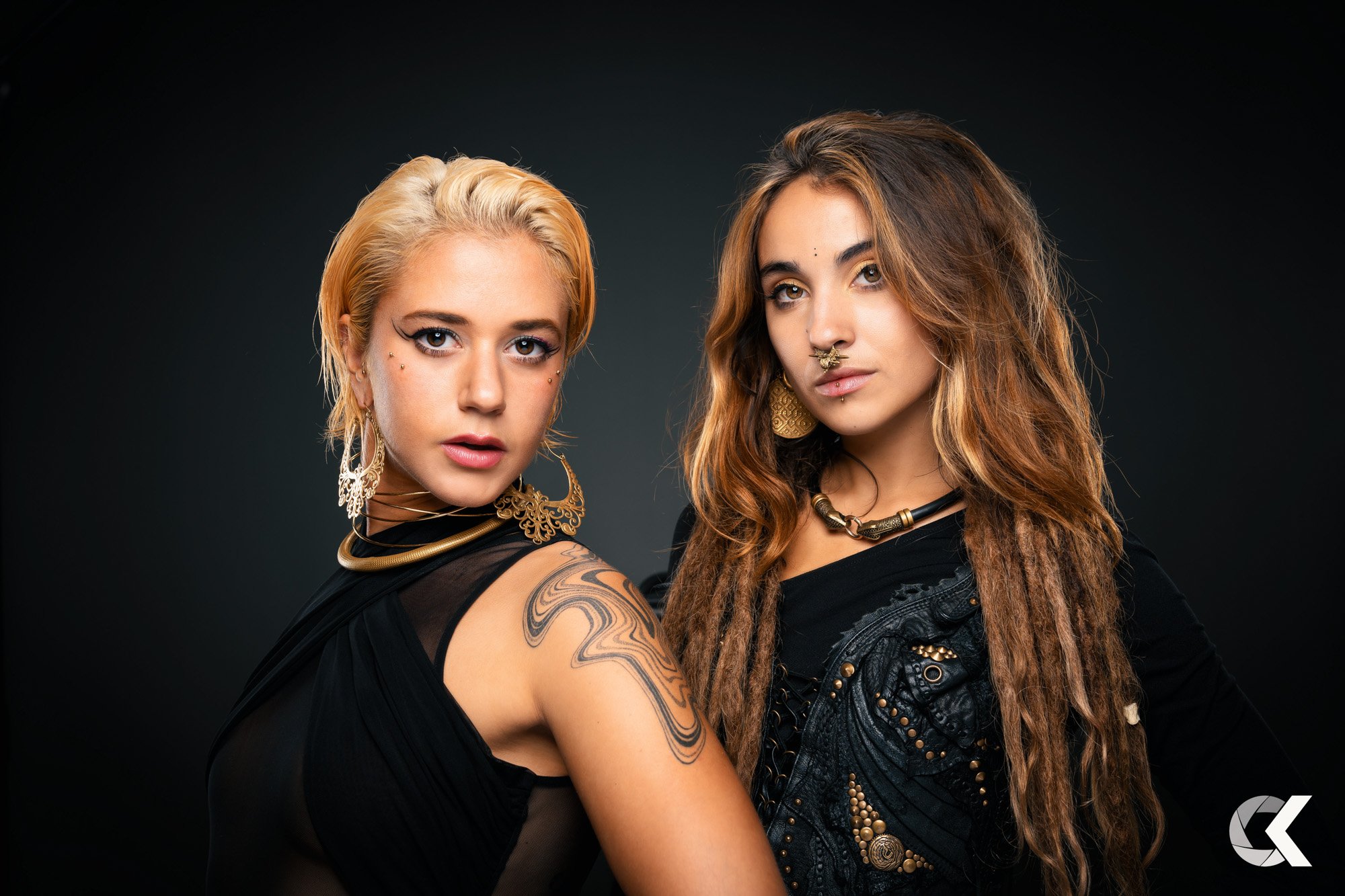 Two women with distinctive jewelry and accessories posing against a dark background, one with blonde hair and tattoos, the other with brown dreadlocks and septum piercing.